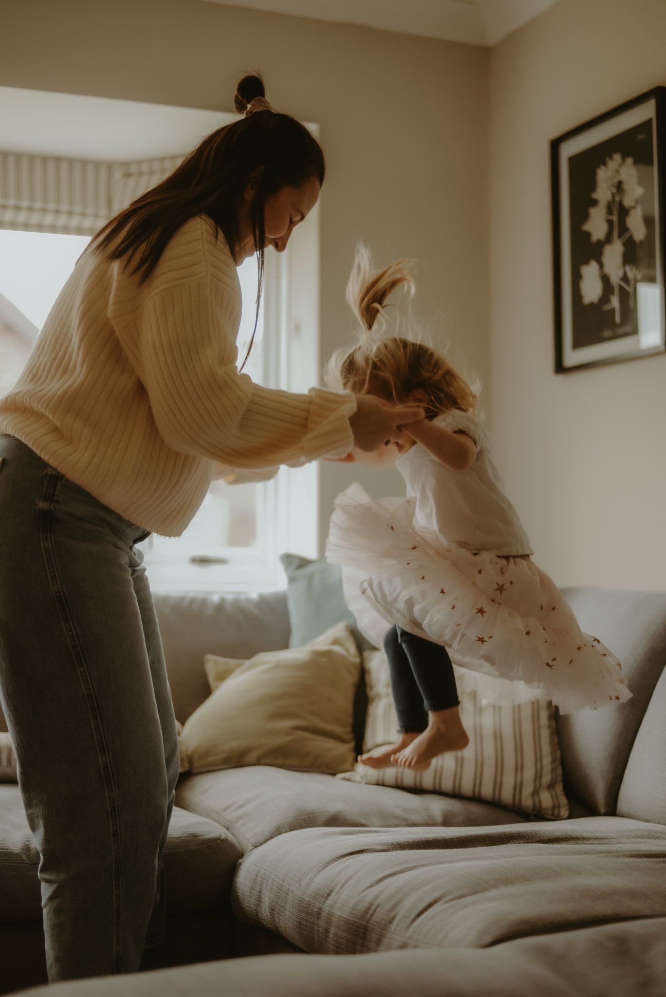 Mother in cream sweater playfully lifts laughing child jumping on a cozy living room sofa.