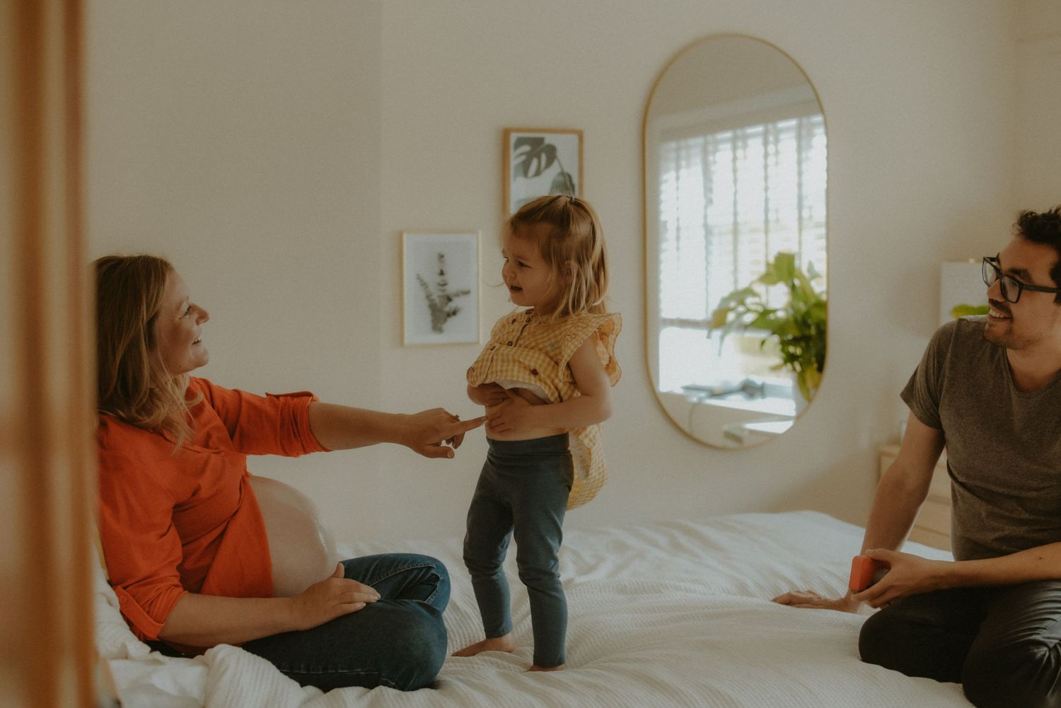 Pregnant woman in orange top sitting on bed with toddler and partner enjoying family playtime together.