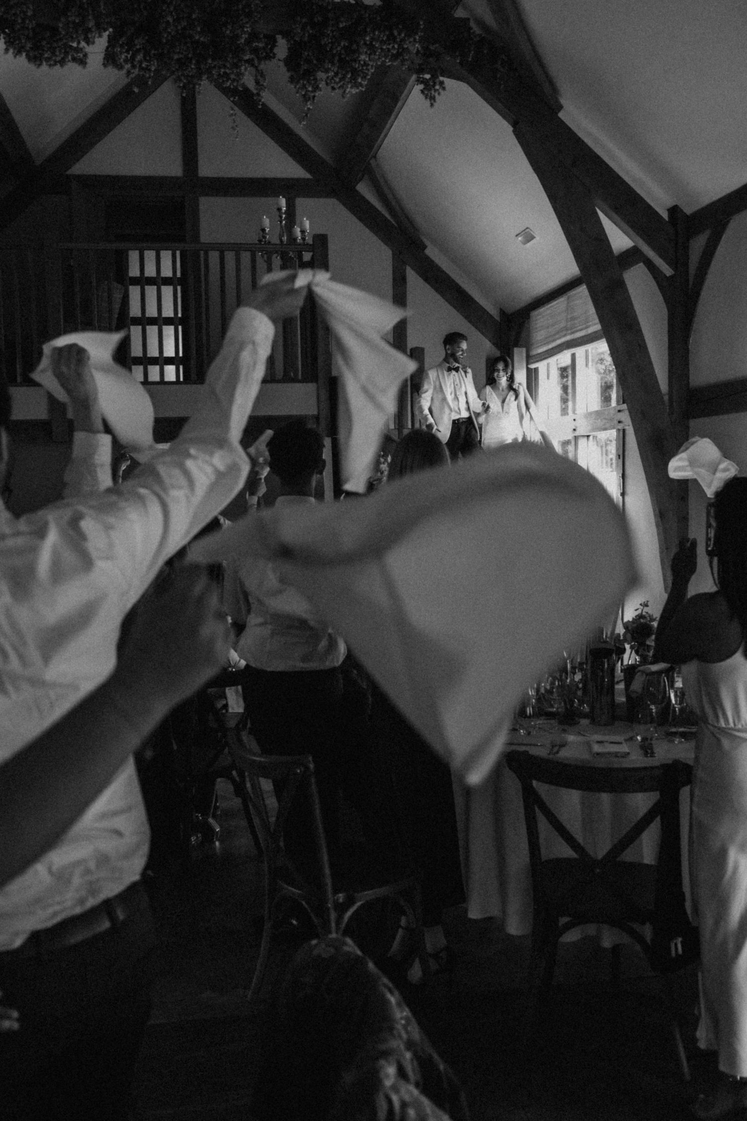 Black and white wedding photo of guests waving white napkins to welcome the newlyweds entering a rustic venue.