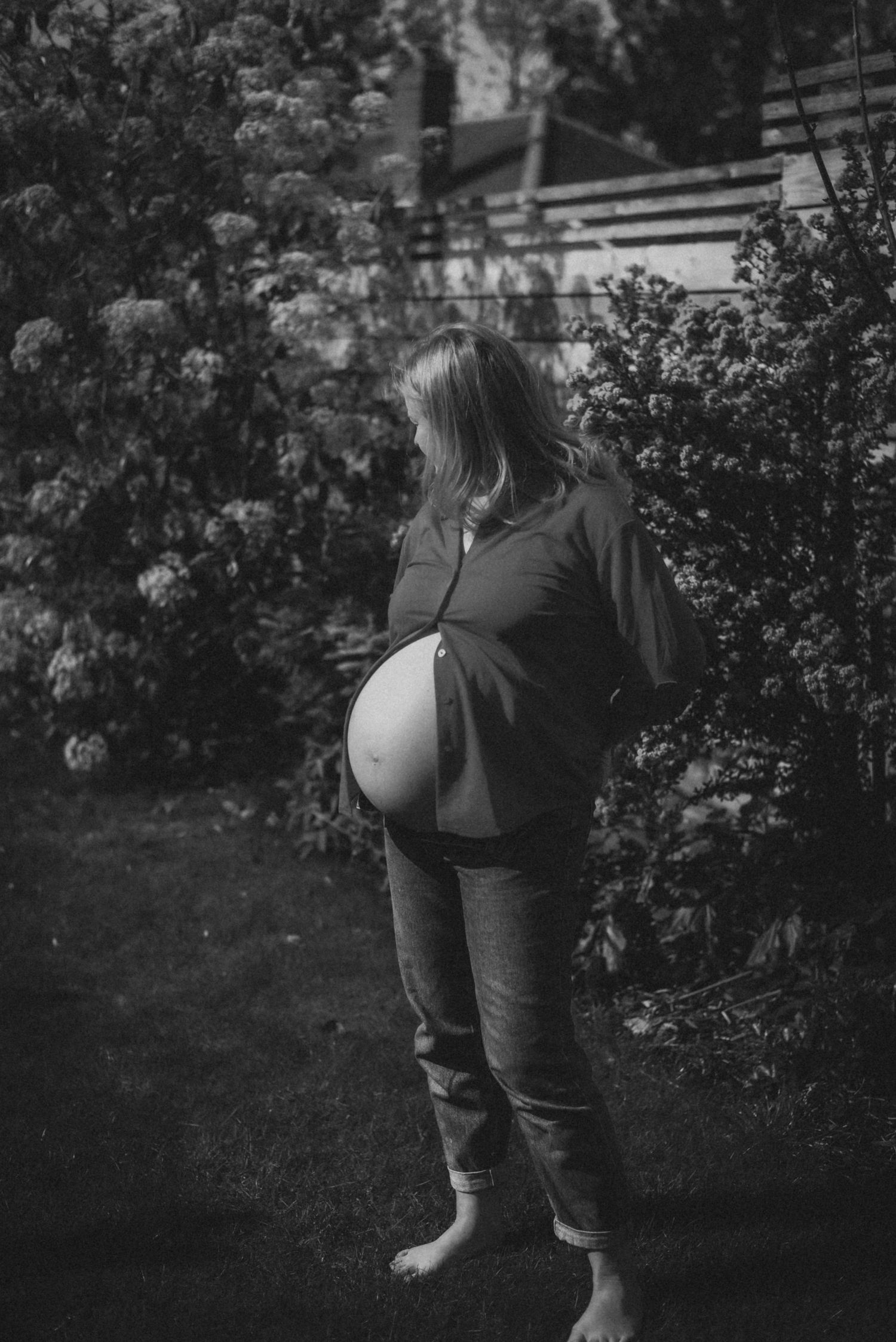Black and white photo of a pregnant woman standing barefoot in a lush garden, her baby bump visible.