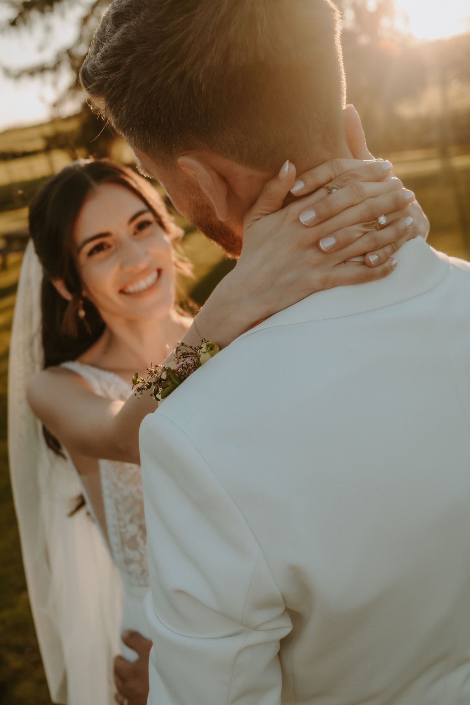 Smiling bride in white dress gazes at groom during golden hour outdoor wedding ceremony.