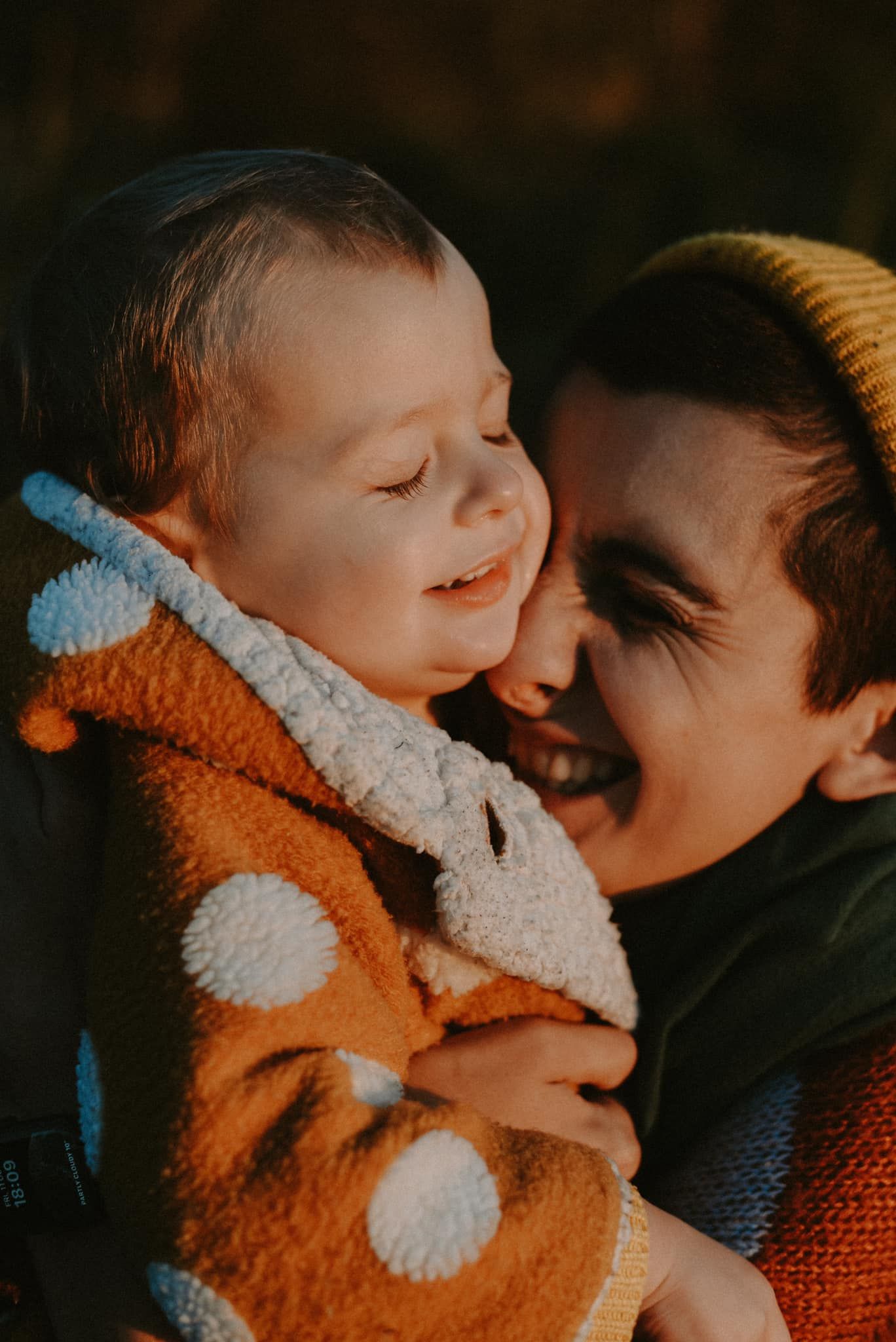 Joyful mother in yellow beanie nuzzling smiling toddler in orange bear jacket outdoors.