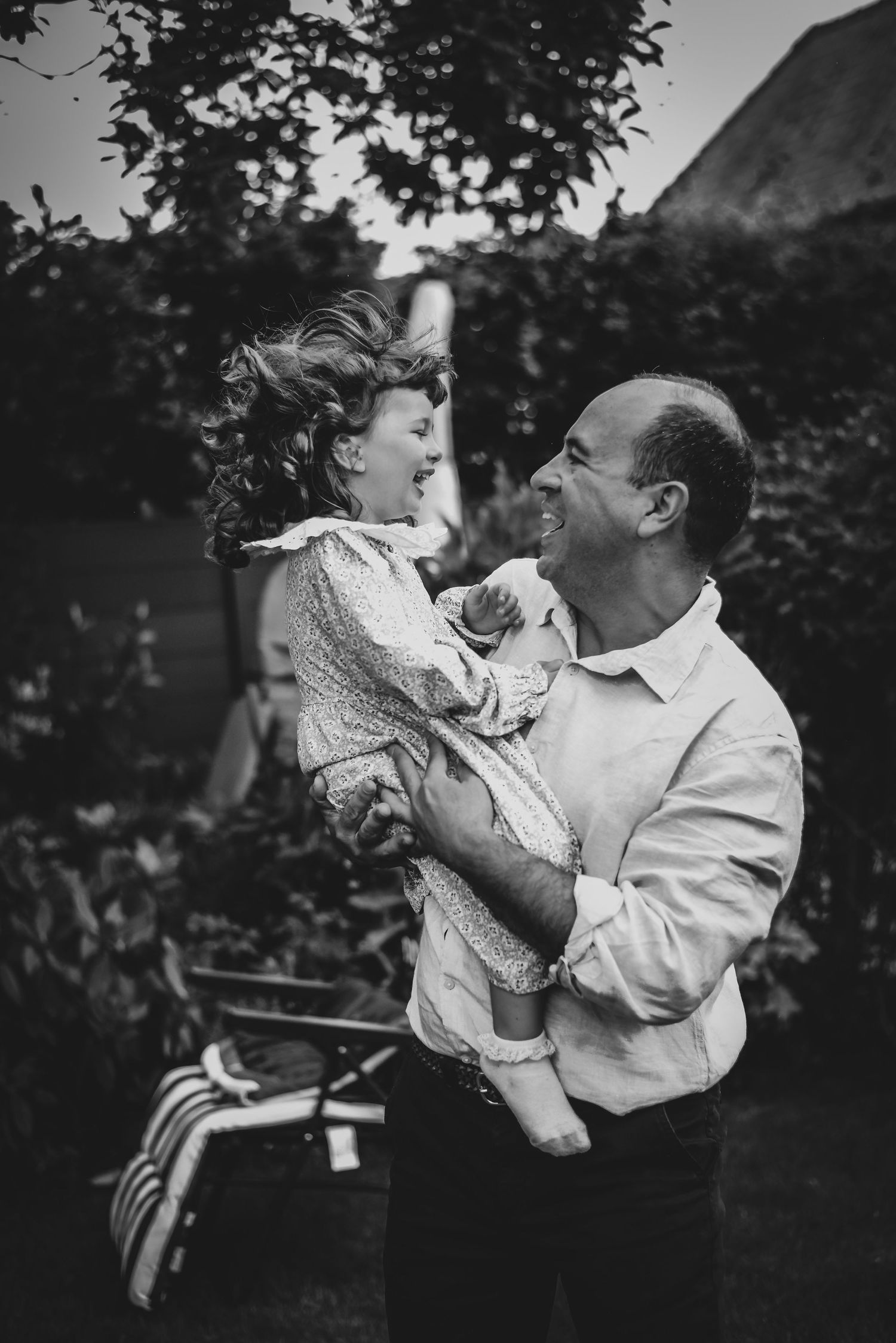 Black and white photo of a laughing father lifting his curly-haired child joyfully in a garden.