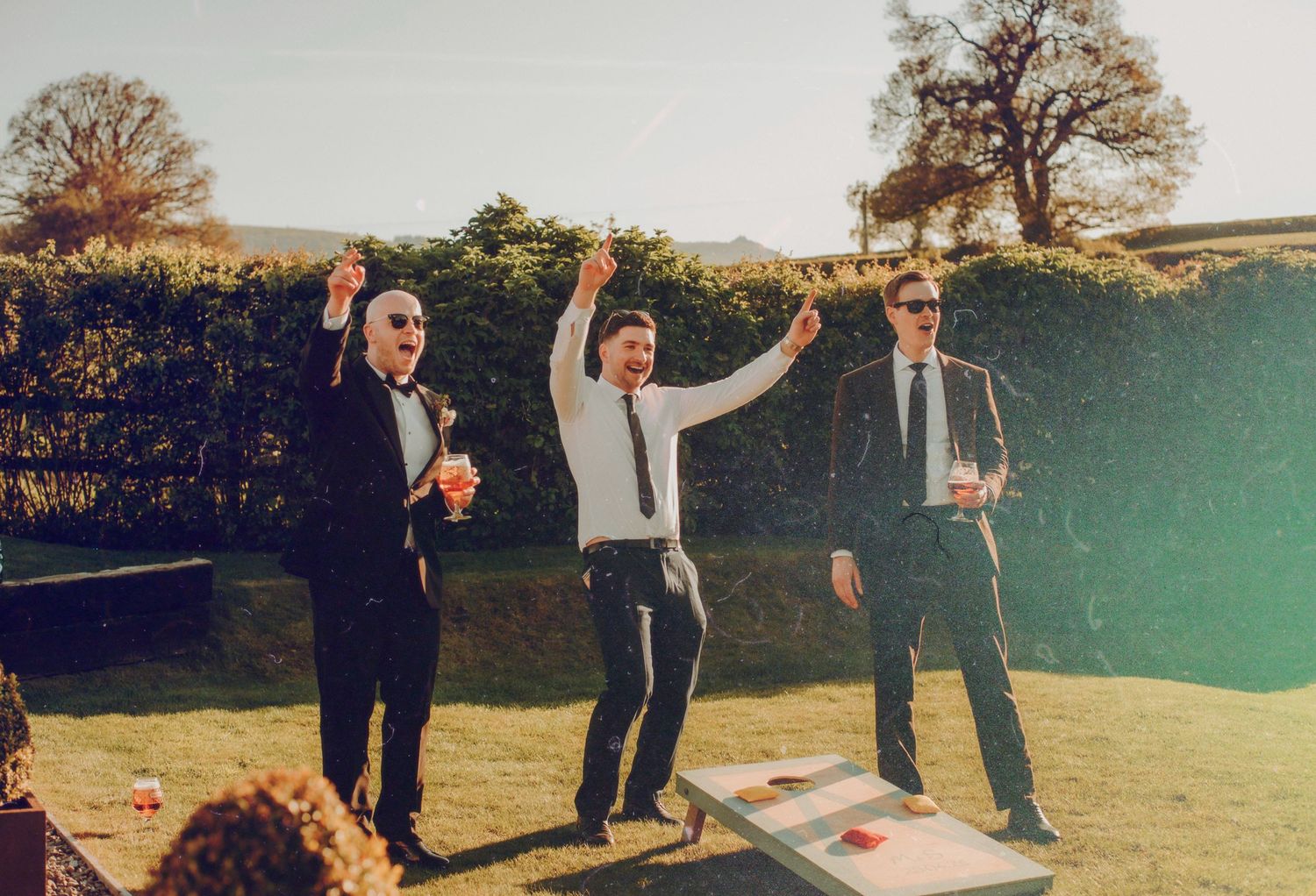 Three men celebrating outdoors at a wedding reception while playing cornhole on a sunny day with a green hedge backdrop.