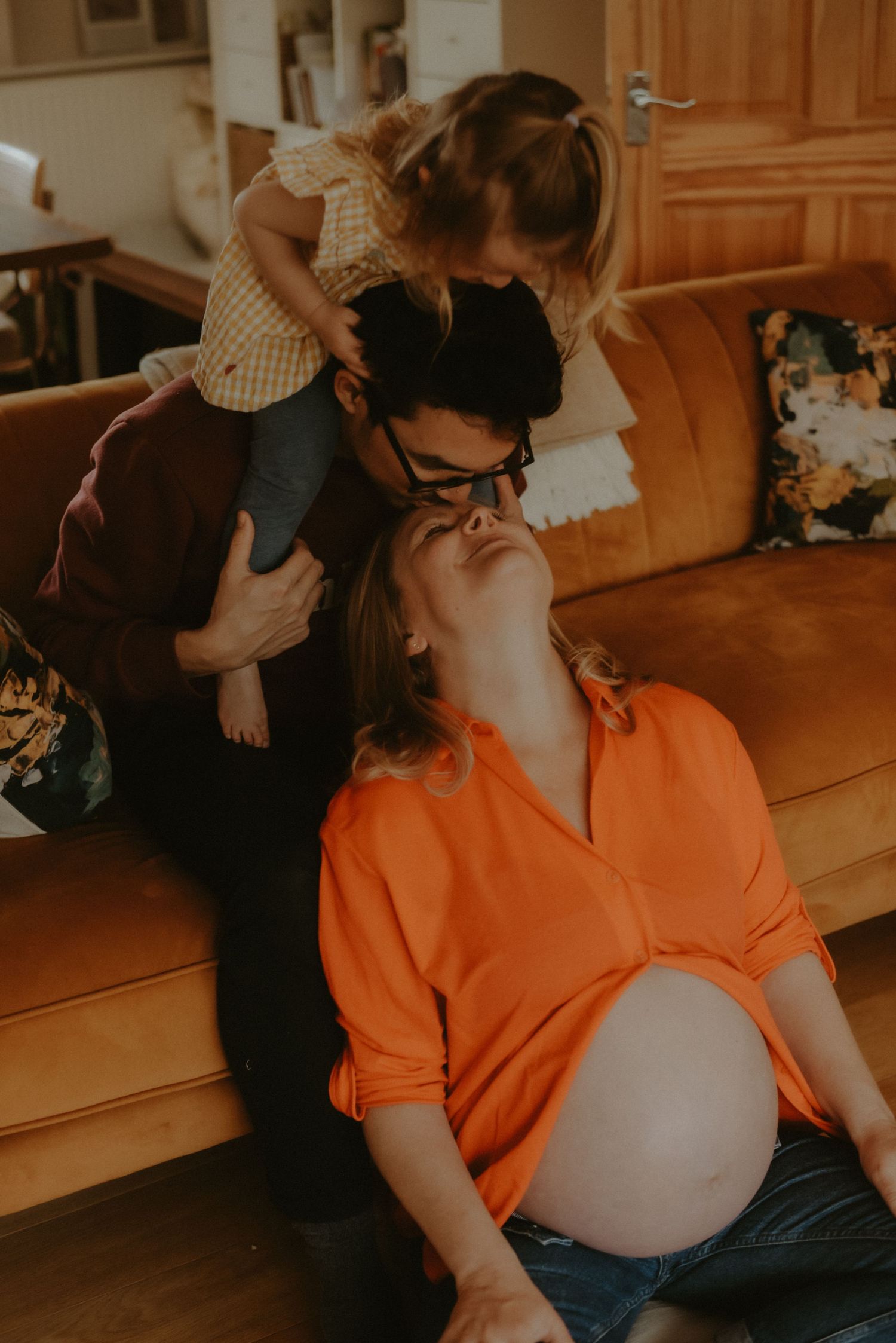 Pregnant woman in orange shirt sits on floor as partner holds toddler girl on couch in warm family moment.