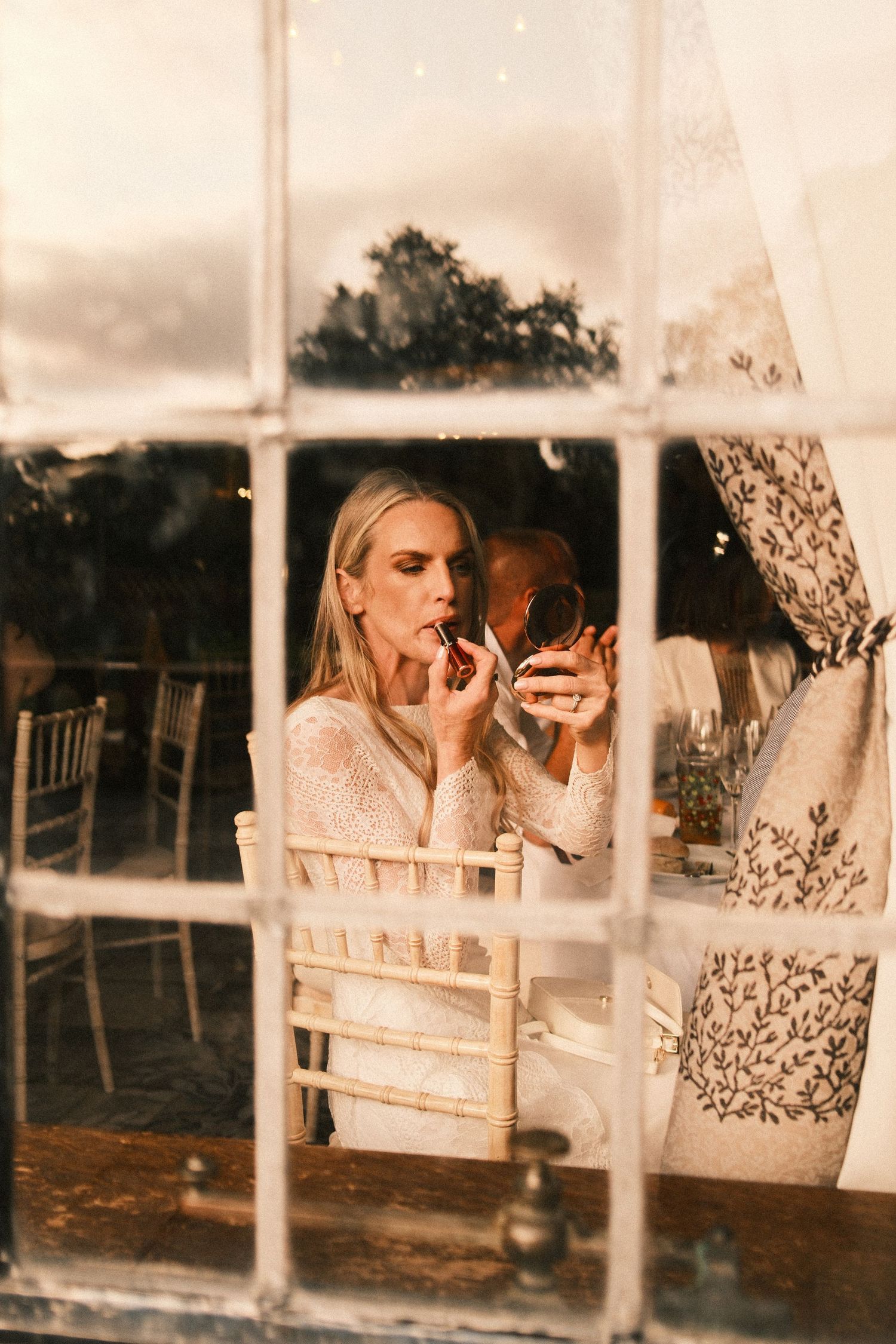 Blonde bride applying lipstick using a compact mirror while seated at a decorated wedding reception table.