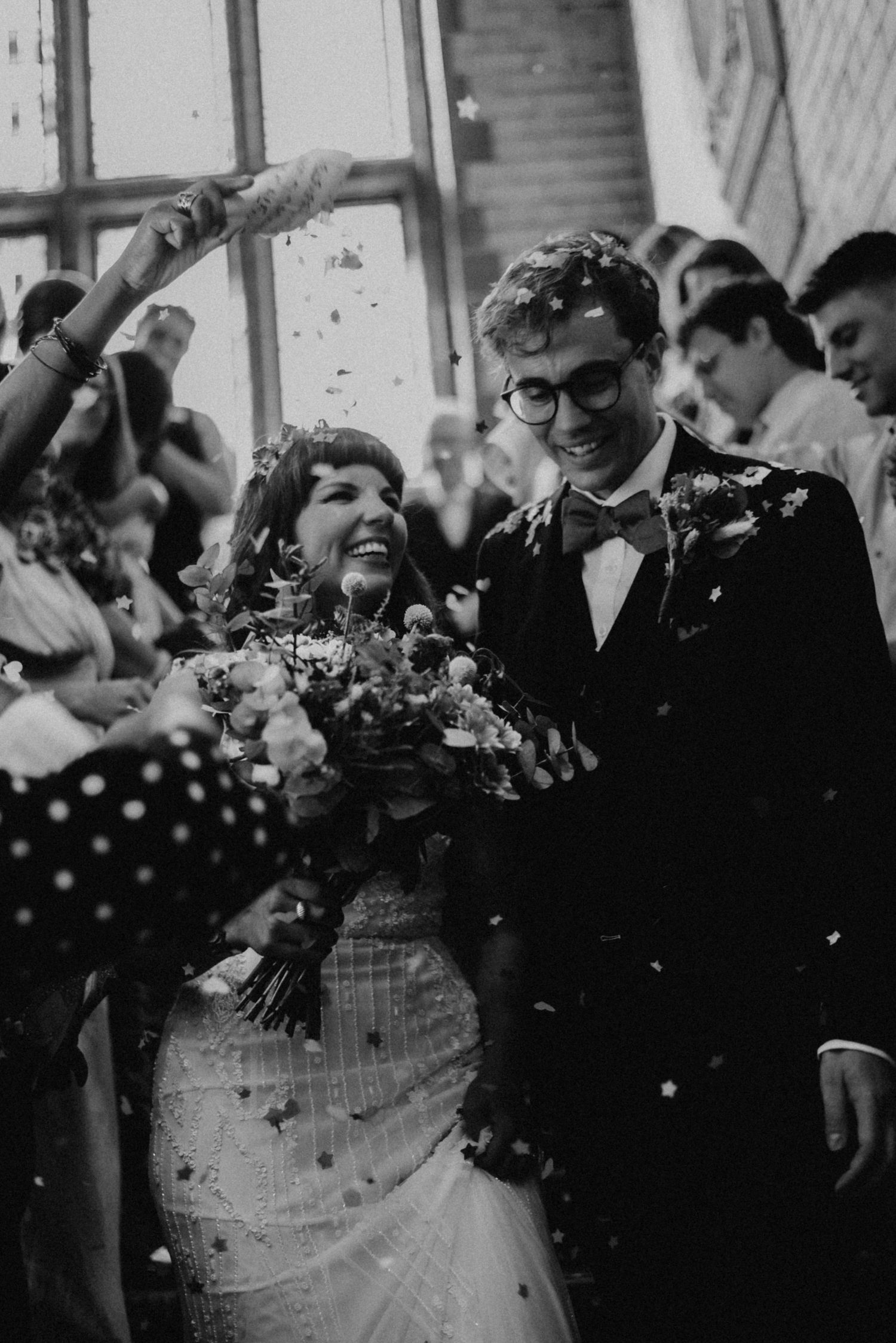 Black and white photo of a joyful bride and groom walking through confetti shower surrounded by cheering wedding guests.