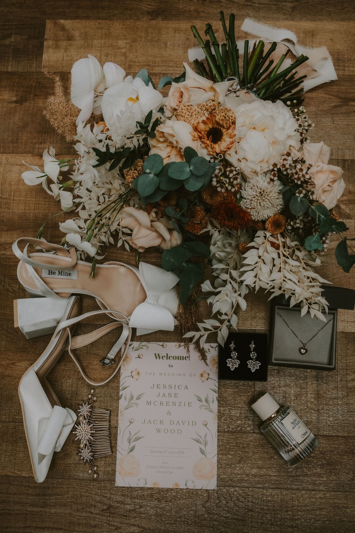 Elegant wedding flat lay featuring bridal bouquet, white heels, jewelry, and ceremony program on rustic wood floor.
