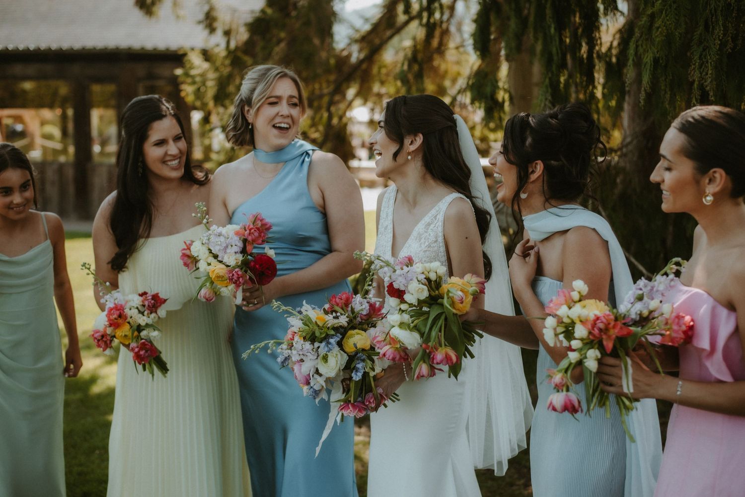 Bridesmaids in pastel dresses laughing and holding colorful bouquets at an outdoor wedding celebration.
