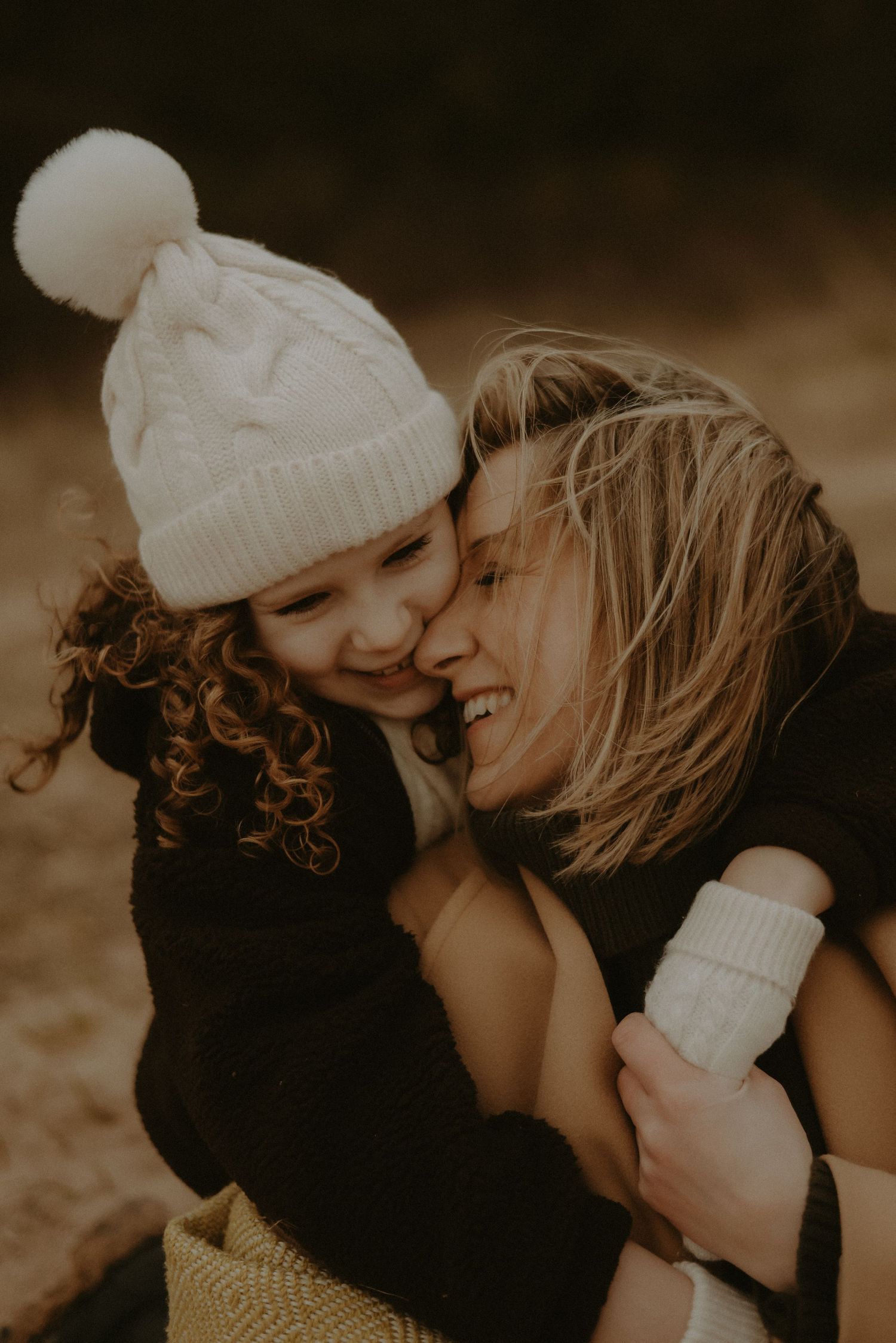 A mother and young daughter share a joyful embrace outdoors, the child wearing a white cable-knit pom-pom hat.