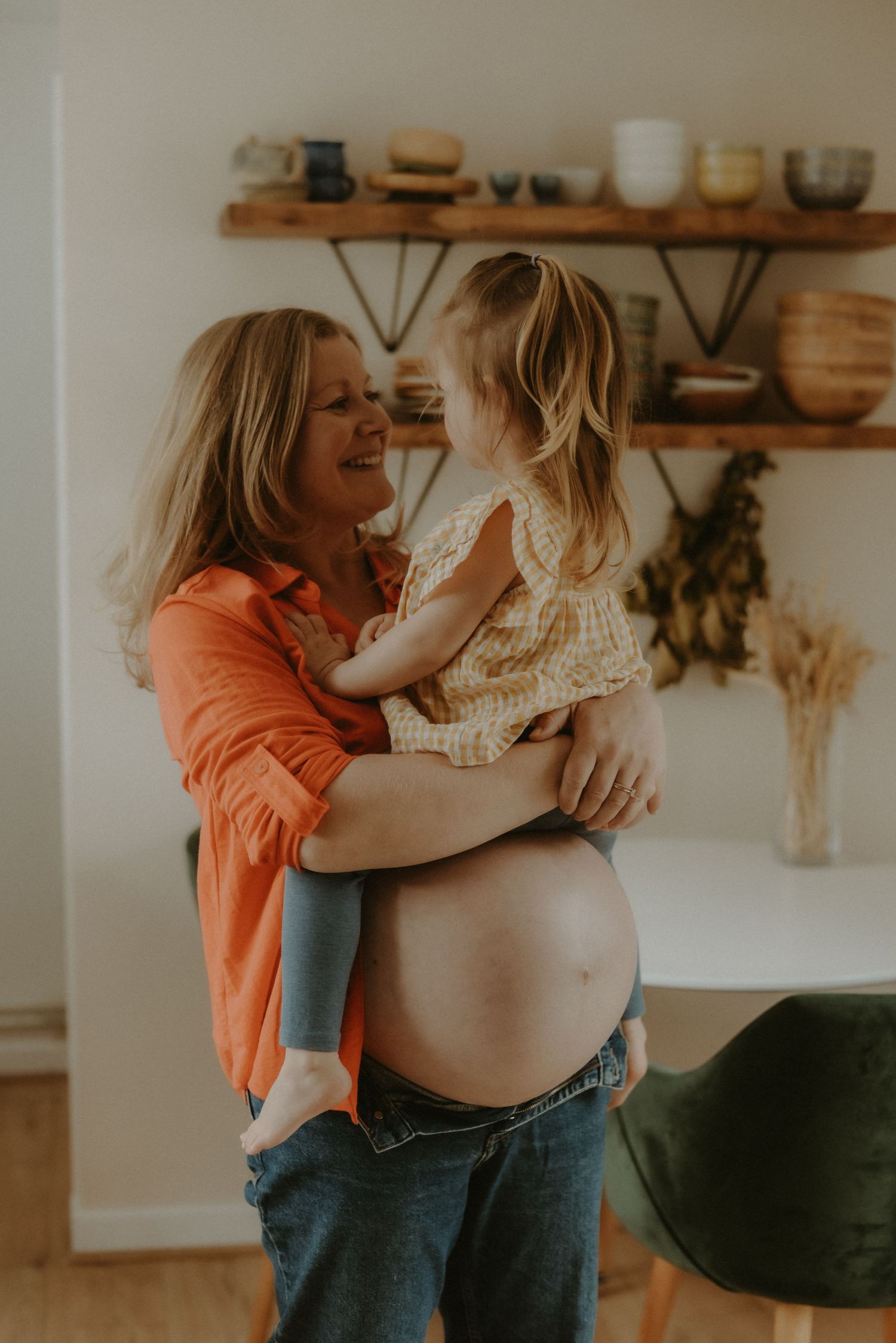 A smiling pregnant woman in an orange shirt lovingly holds a young toddler girl in her arms.