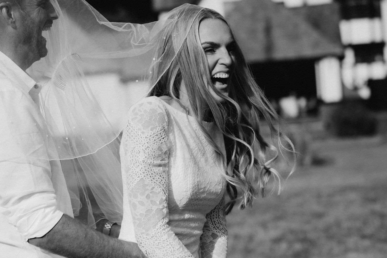 Black and white photo of a laughing bride in lace dress with veil blowing in wind on her wedding day.