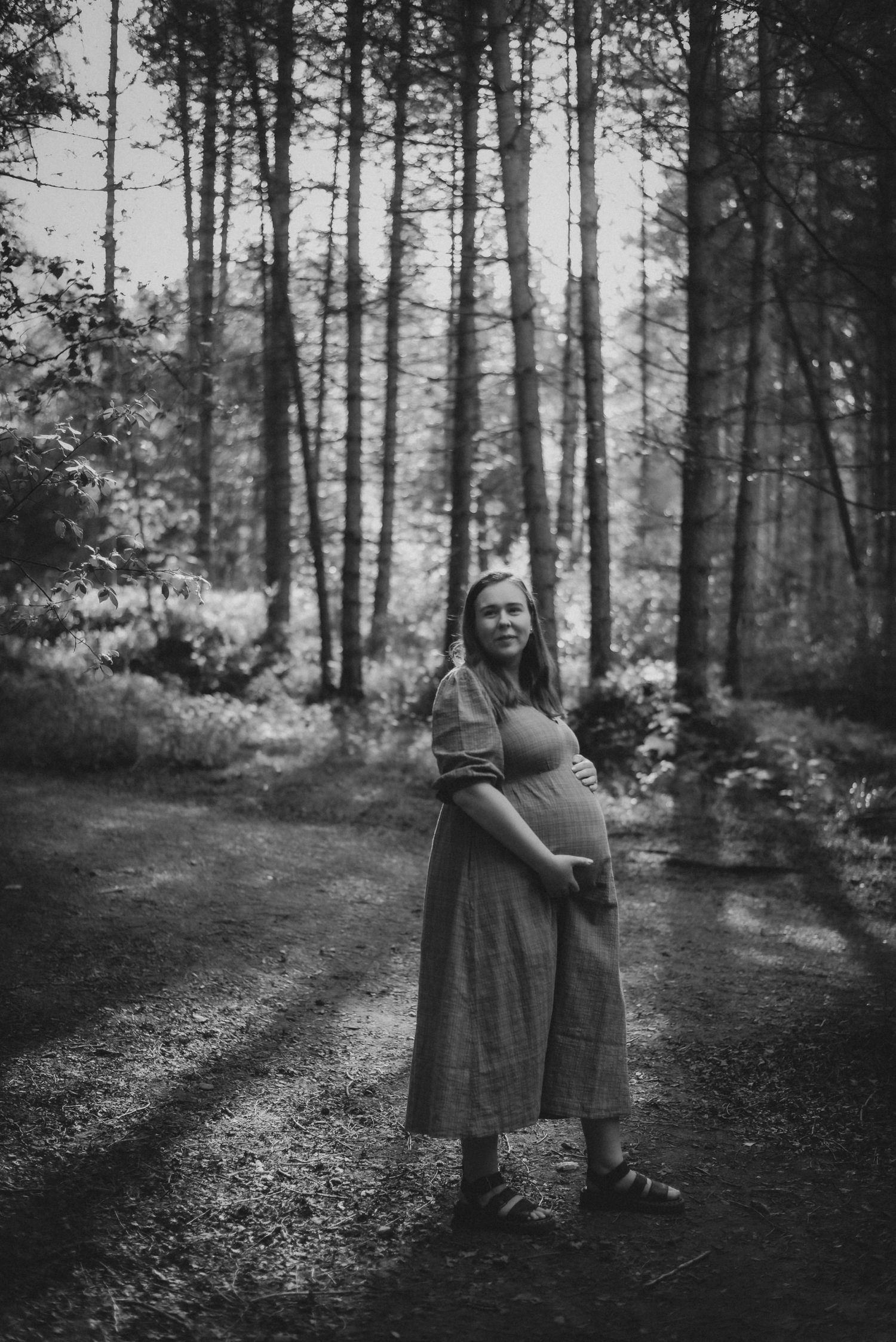 Black and white photo of a pregnant woman in a maxi dress standing on a forest path, cradling her baby bump.