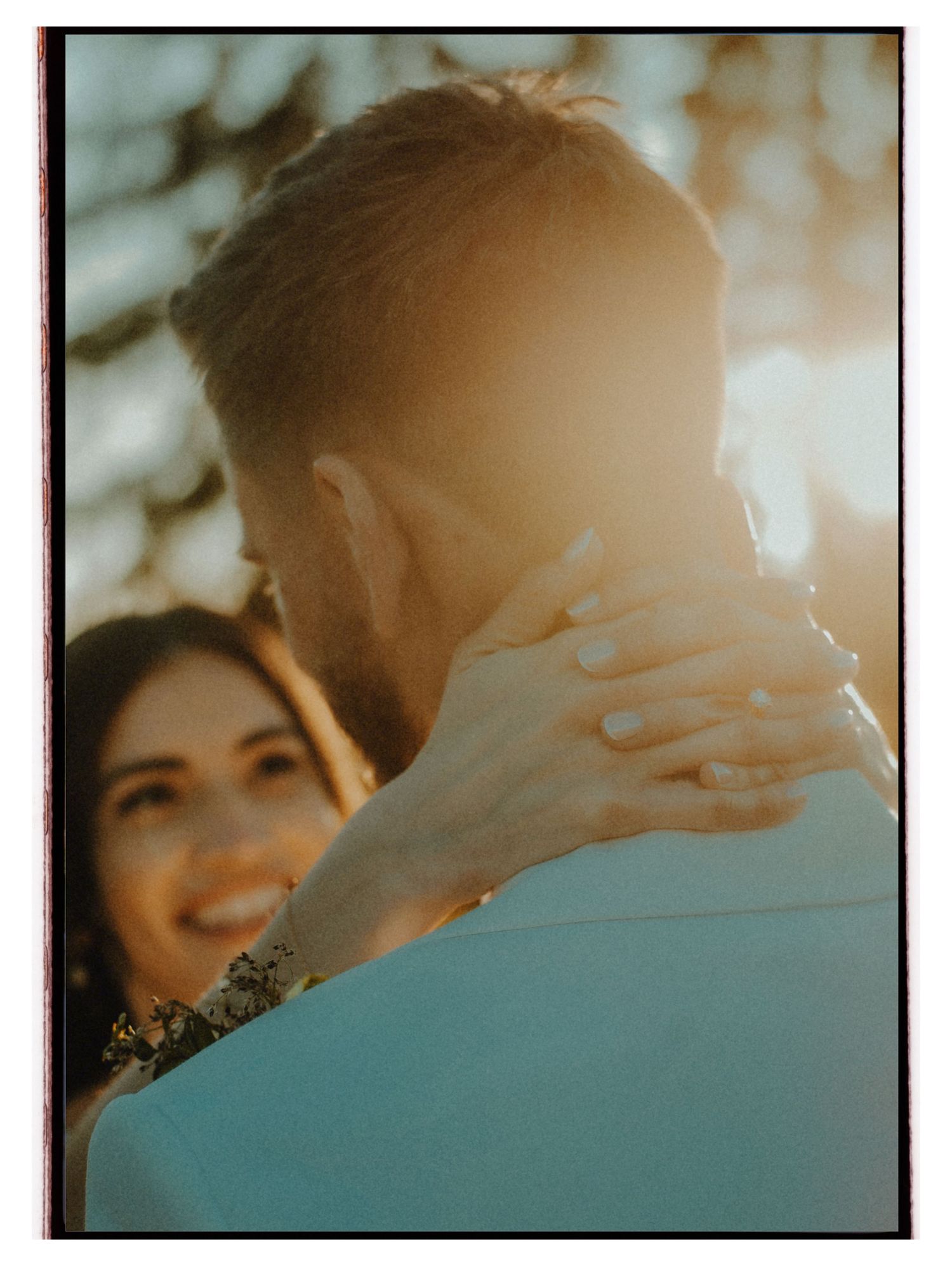 Couple embracing outdoors at golden hour, woman smiling with hand on man's neck, warm sunlight glowing behind them.