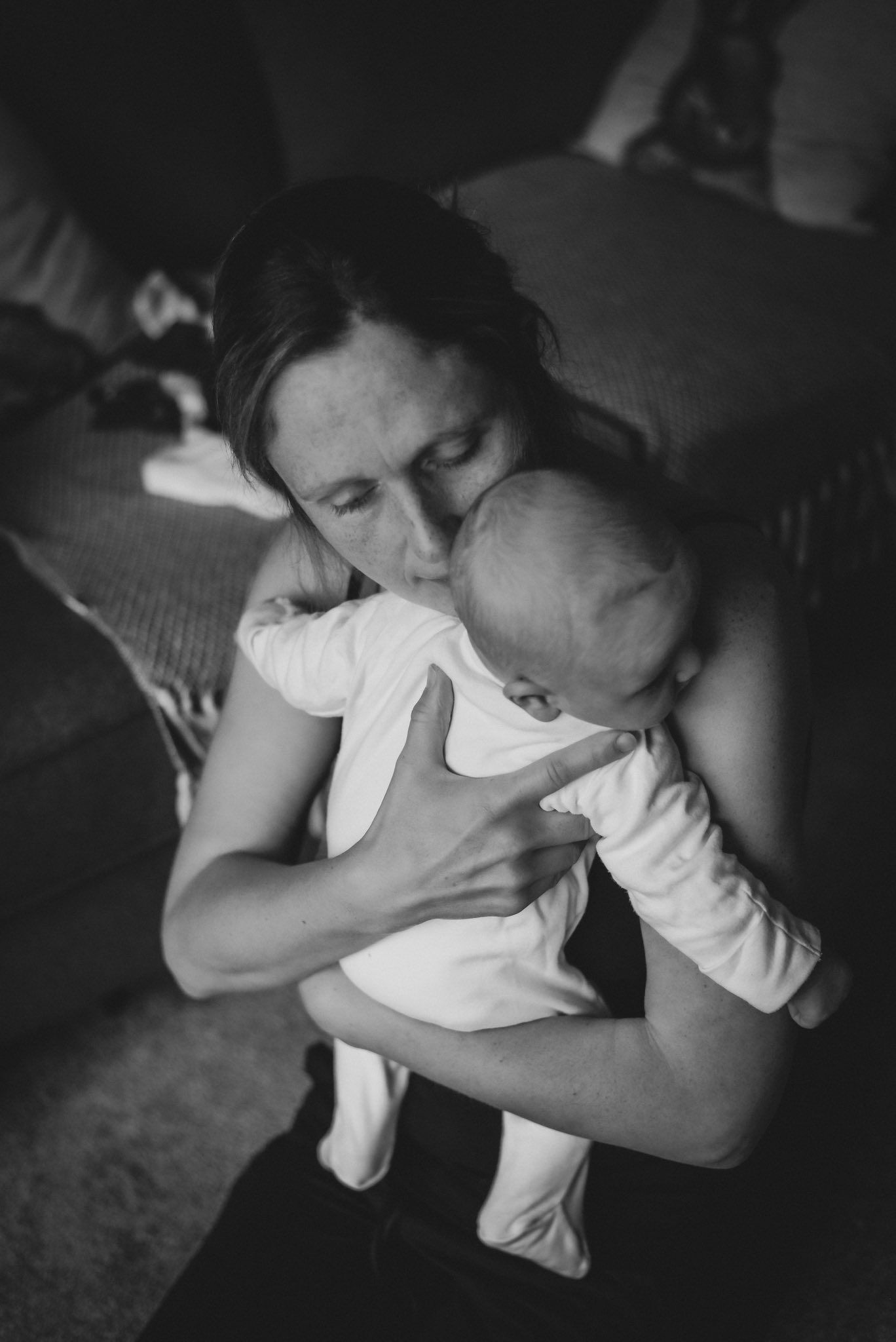 Black and white photo of a mother tenderly kissing her newborn baby indoors.