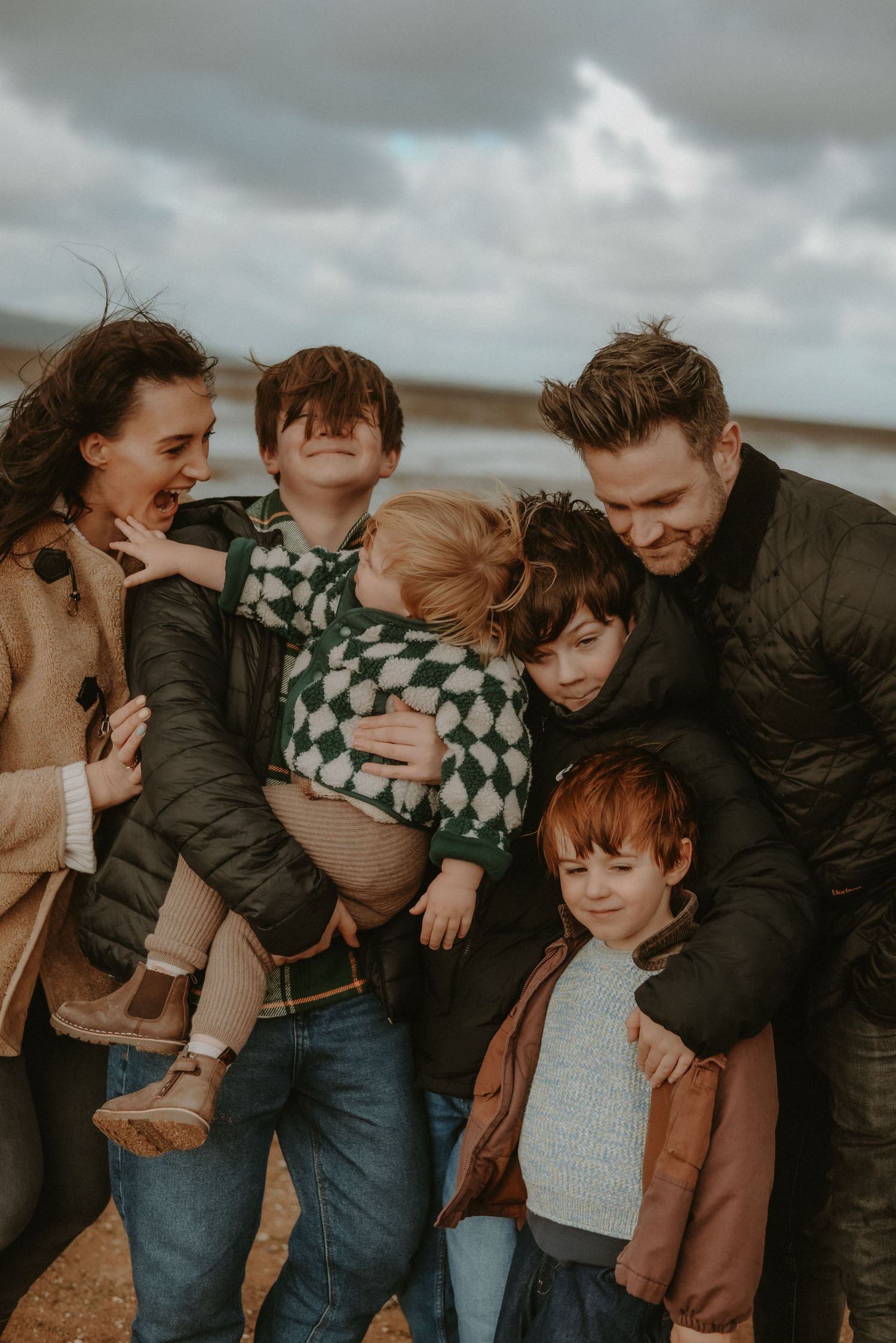 Family of six laughing and hugging outdoors on a cloudy beach day, parents embracing children in casual winter jackets. Thurstaston beach