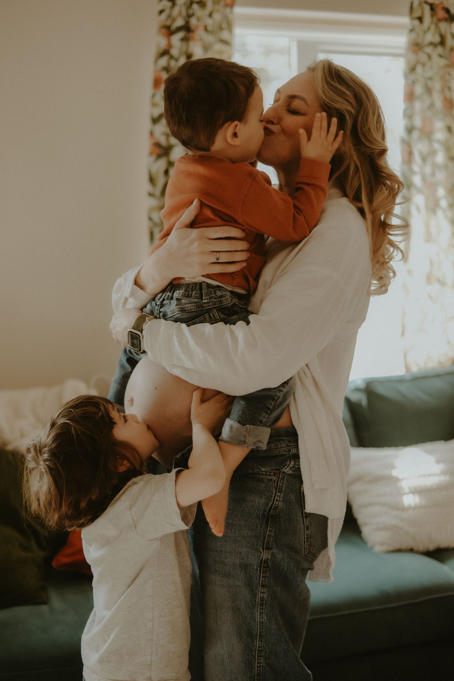 A mother kisses her toddler while her young child hugs her legs in a warm, sunlit living room.