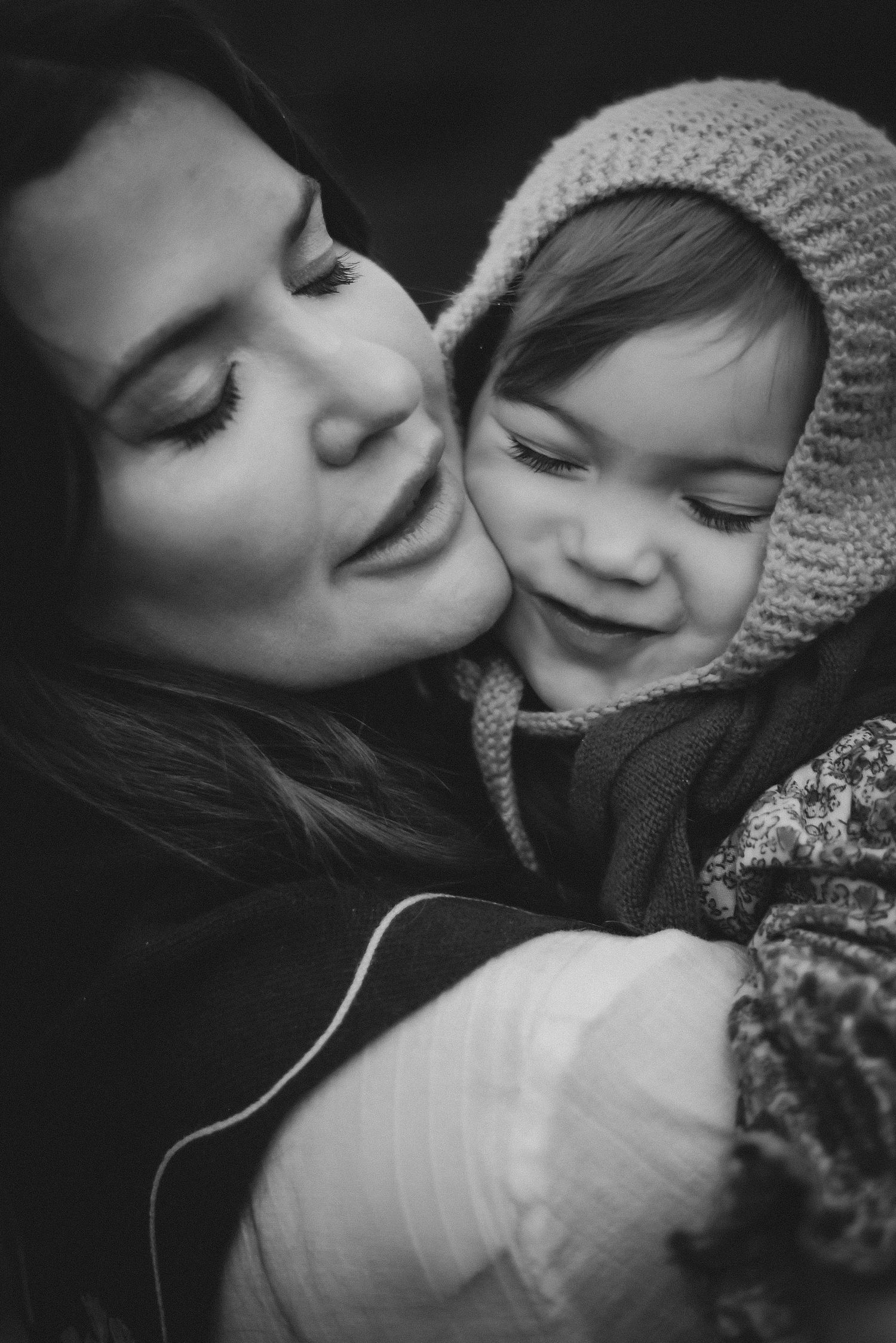 Black and white close-up of a mother tenderly kissing her smiling baby in a knit bonnet.