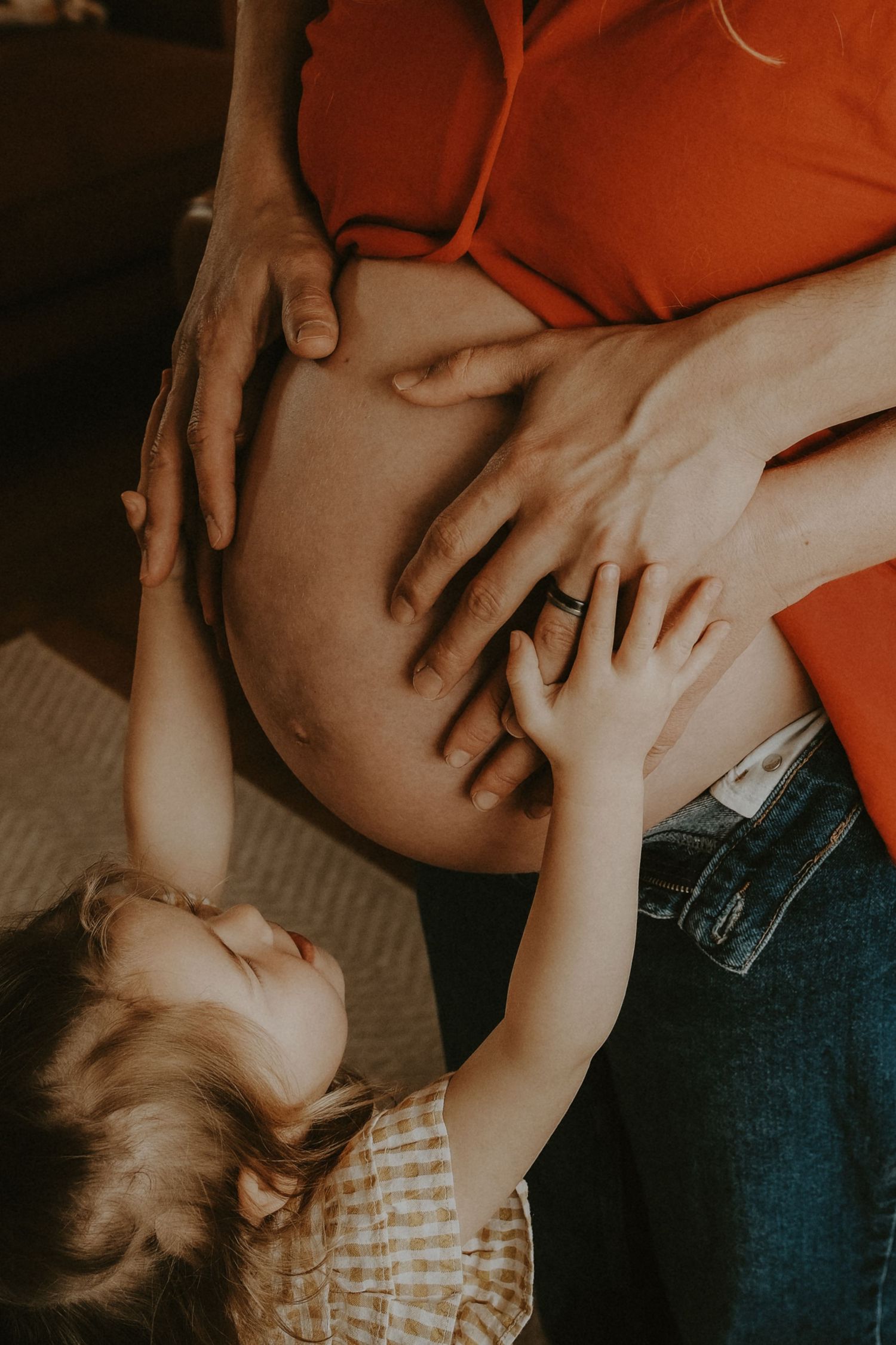 A toddler touching a pregnant woman's bare belly, with adult hands gently resting alongside.