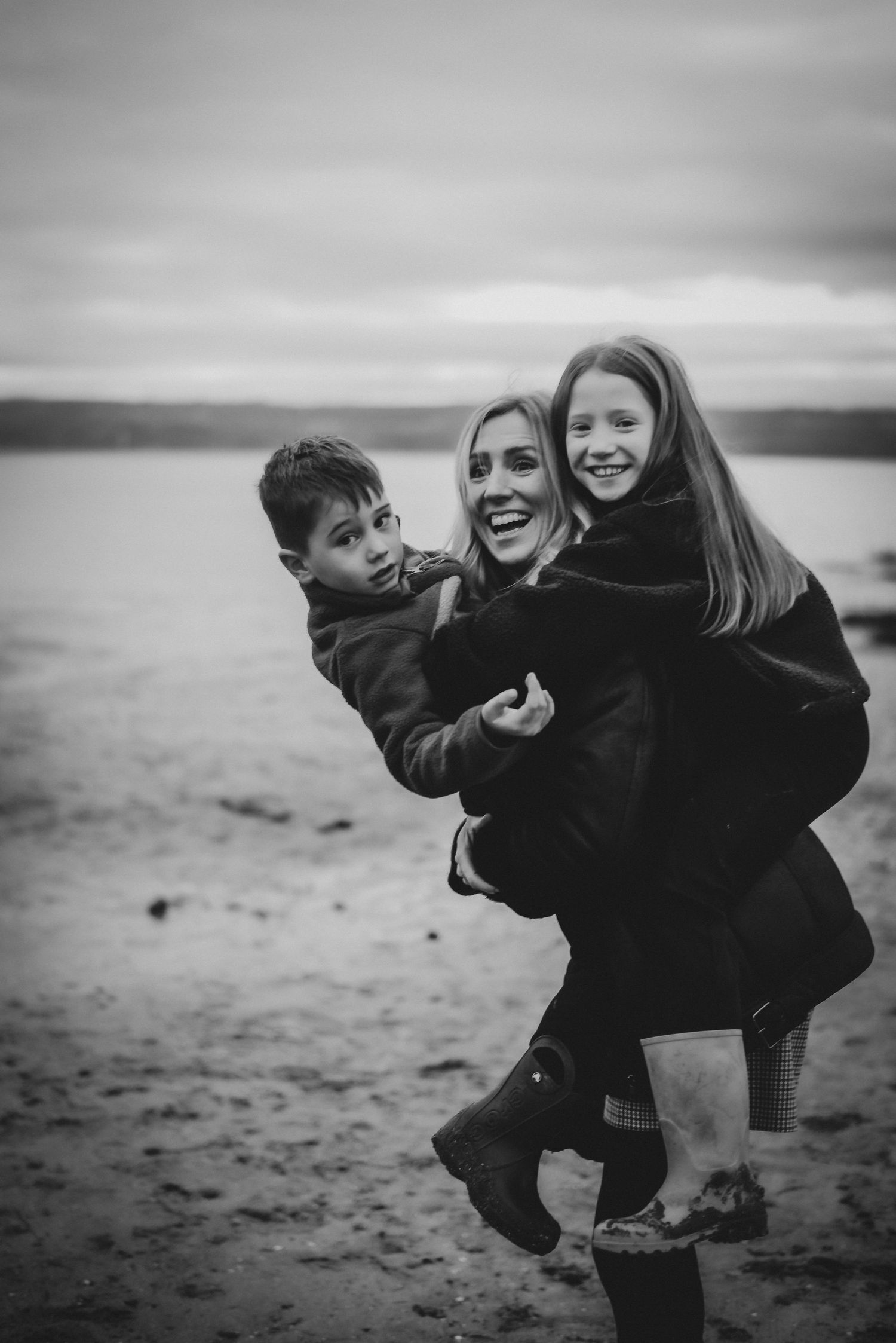Black and white photo of a smiling woman carrying two children on a beach, enjoying a fun family moment outdoors.