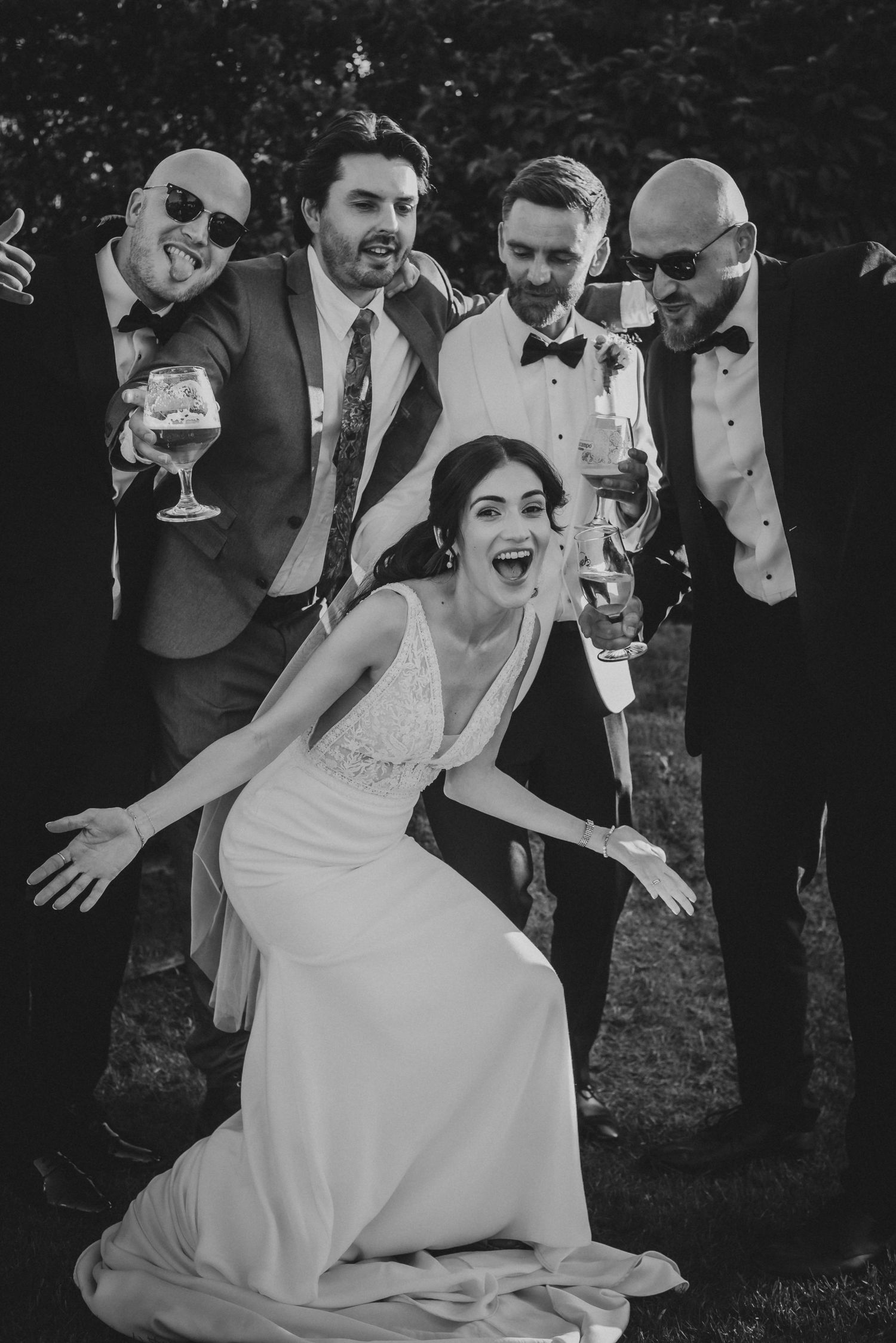 Black and white photo of a joyful bride posing with four groomsmen holding drinks at a wedding celebration.