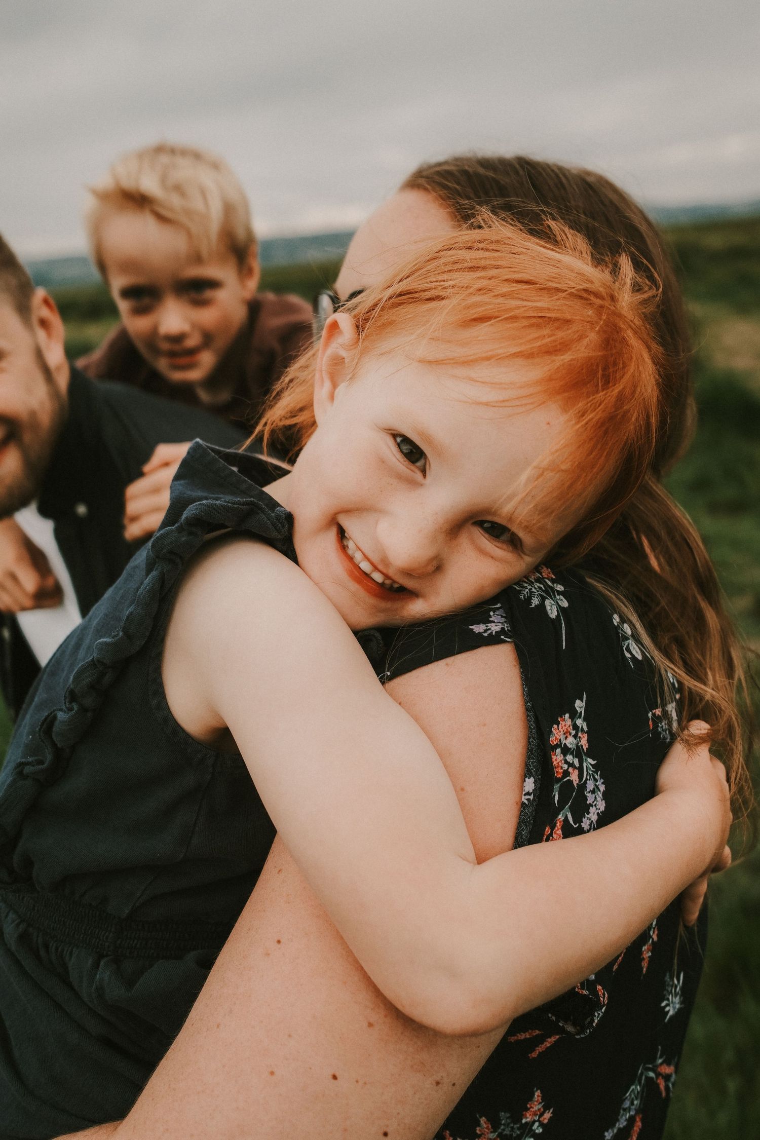 Happy red-haired girl smiling while hugging an adult outdoors with a boy laughing in the background.