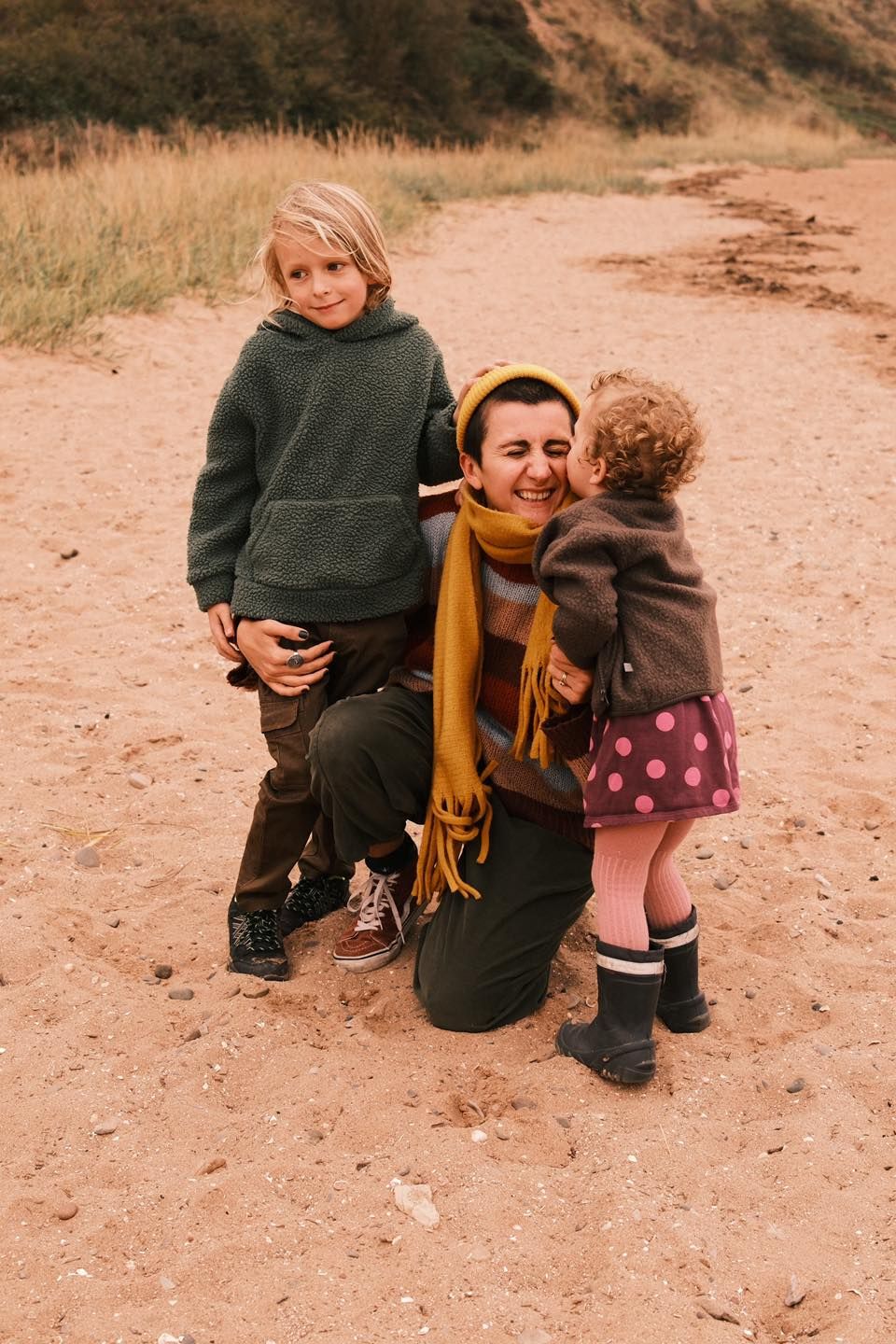 A smiling mother kneels on sandy ground hugging her two young children outdoors.