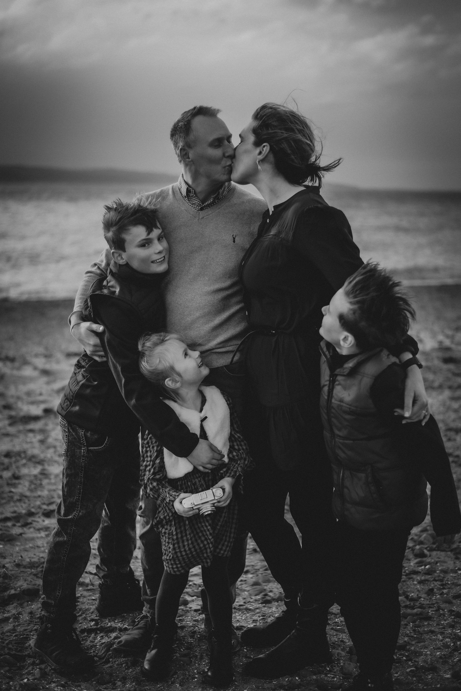 Black and white beach portrait of a family of five with parents kissing and children embracing.