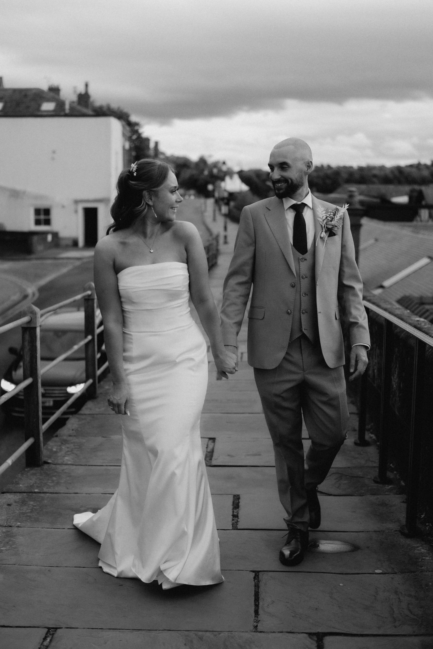 Black and white photo of a bride and groom holding hands, walking together on a stone path on their wedding day.