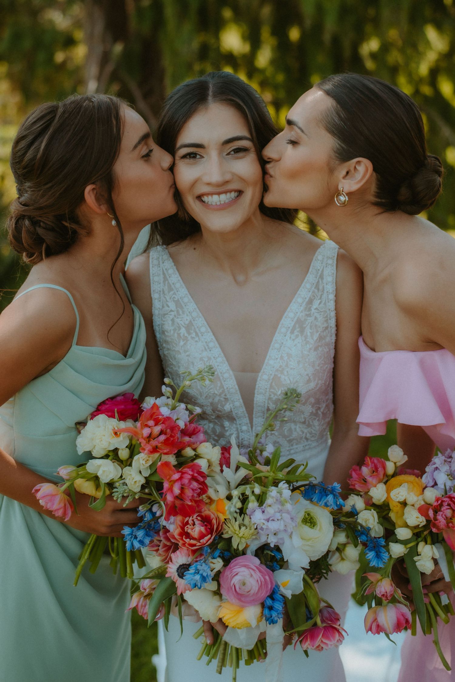 Bride in white lace gown smiles as two bridesmaids in mint and pink dresses kiss her cheeks, holding colorful bouquets.