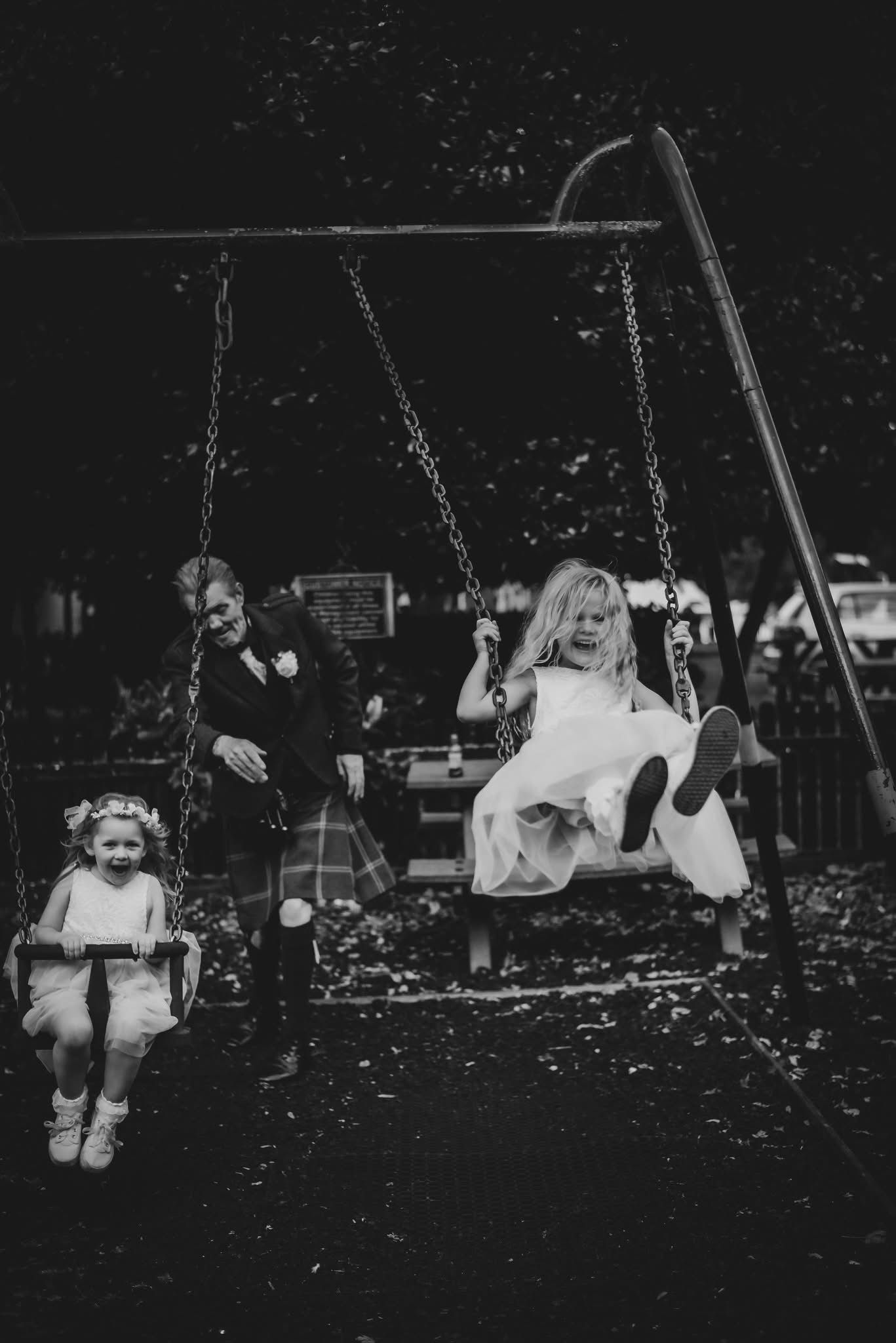 Black and white photo of children in formal attire playing on swings at a wedding celebration outdoors.