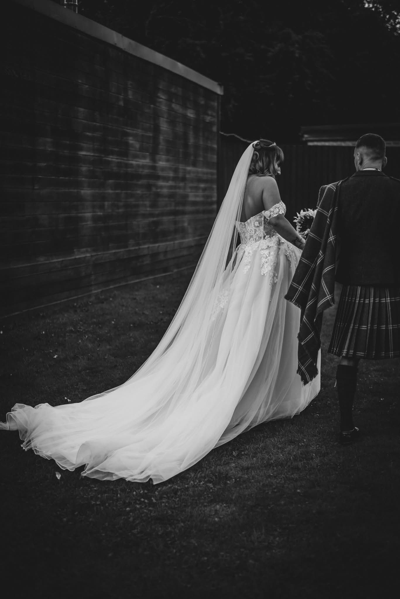 Black and white photo of a bride in a flowing gown and groom in a tartan kilt walking together outdoors.