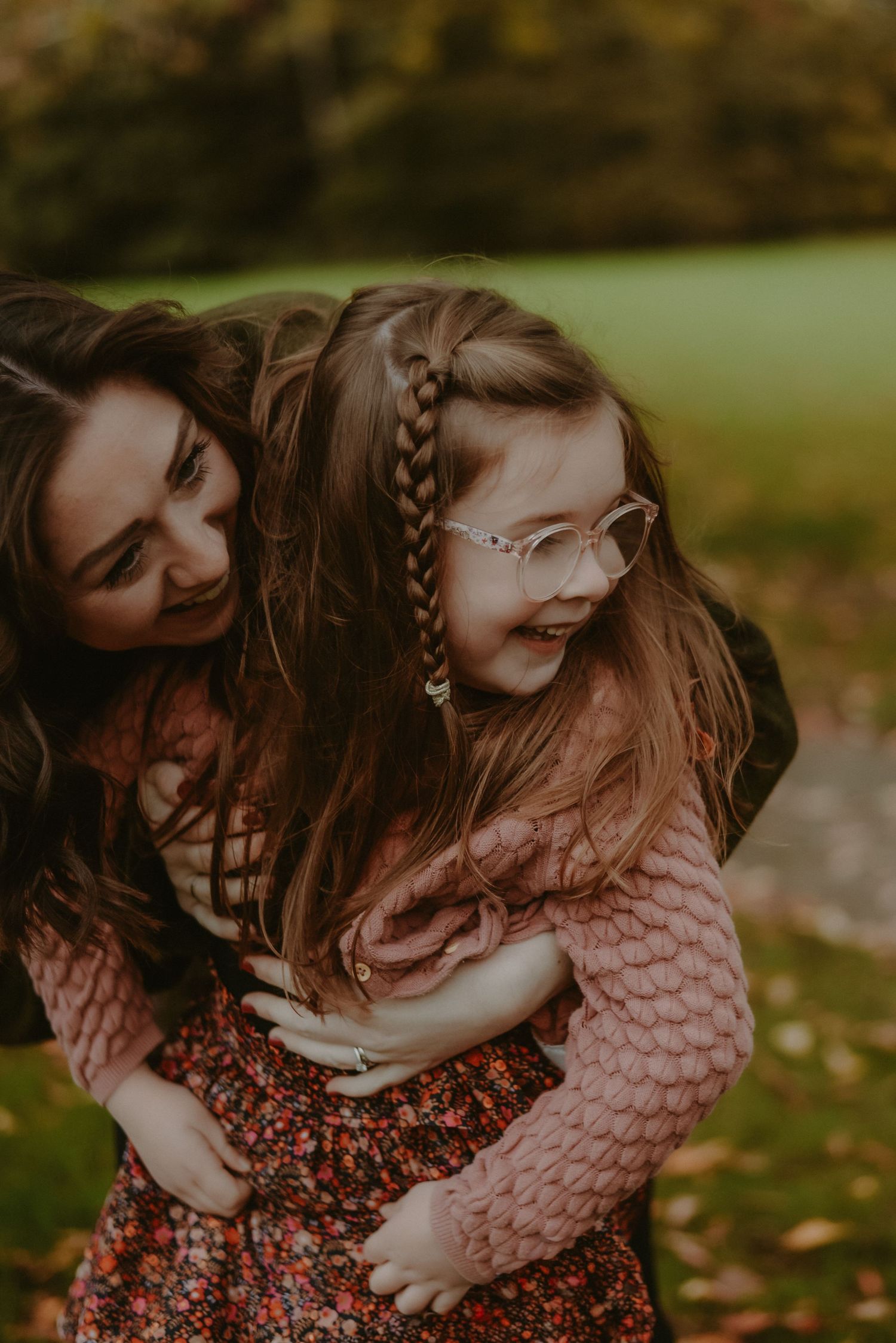 Mother hugging smiling young girl wearing glasses and braids in autumn park.