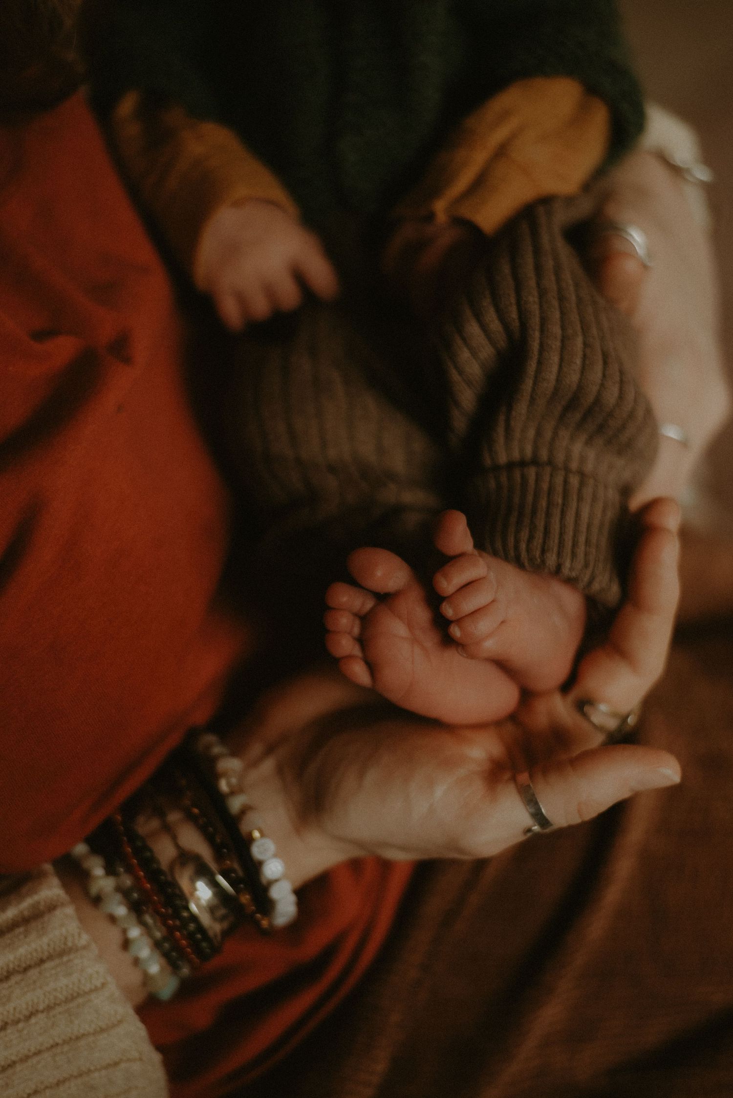 Close-up of newborn baby feet cradled in parent hands with bracelets and rings.