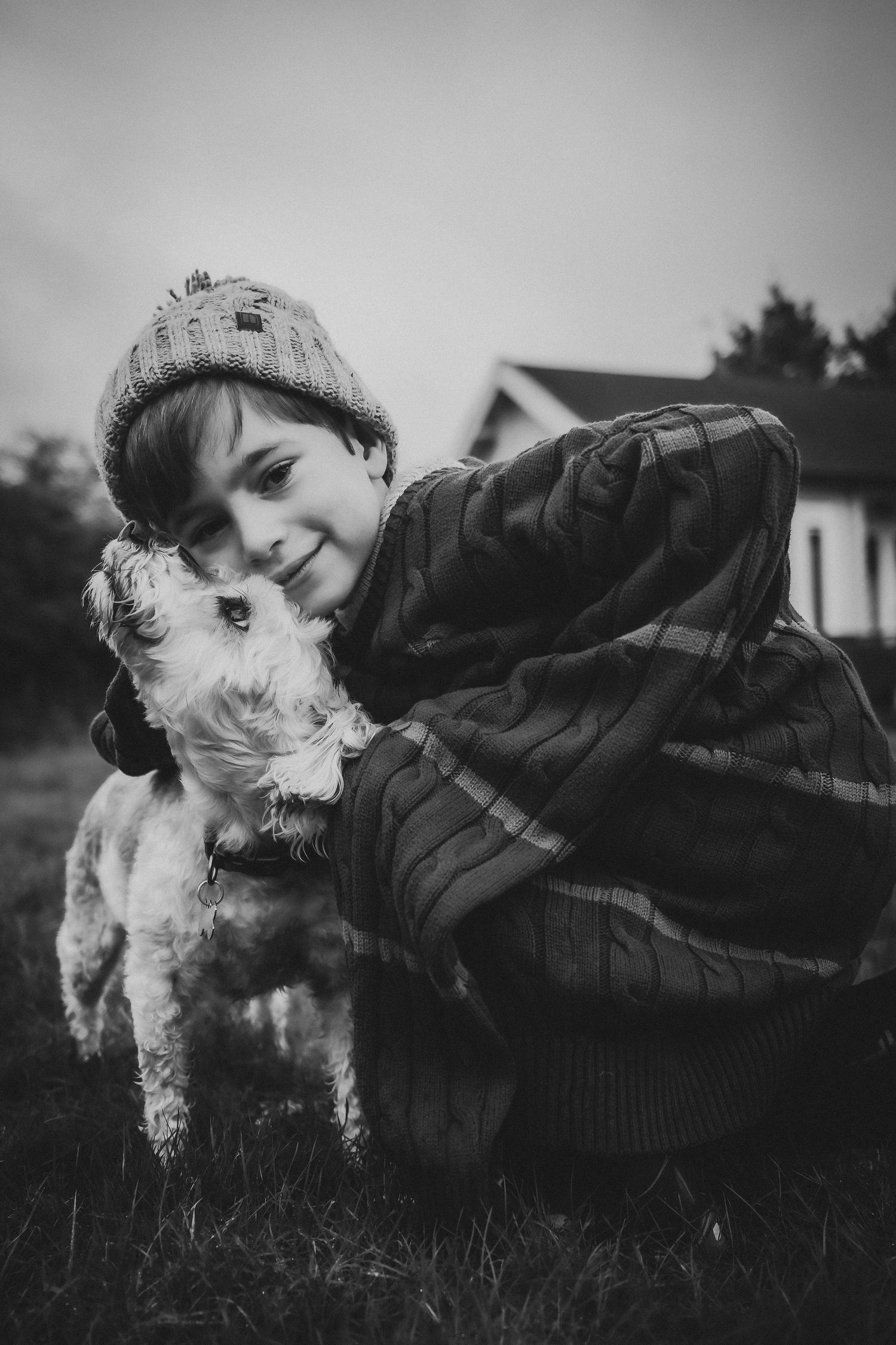 Black and white photo of a smiling boy in a knit hat hugging his fluffy white dog outdoors.