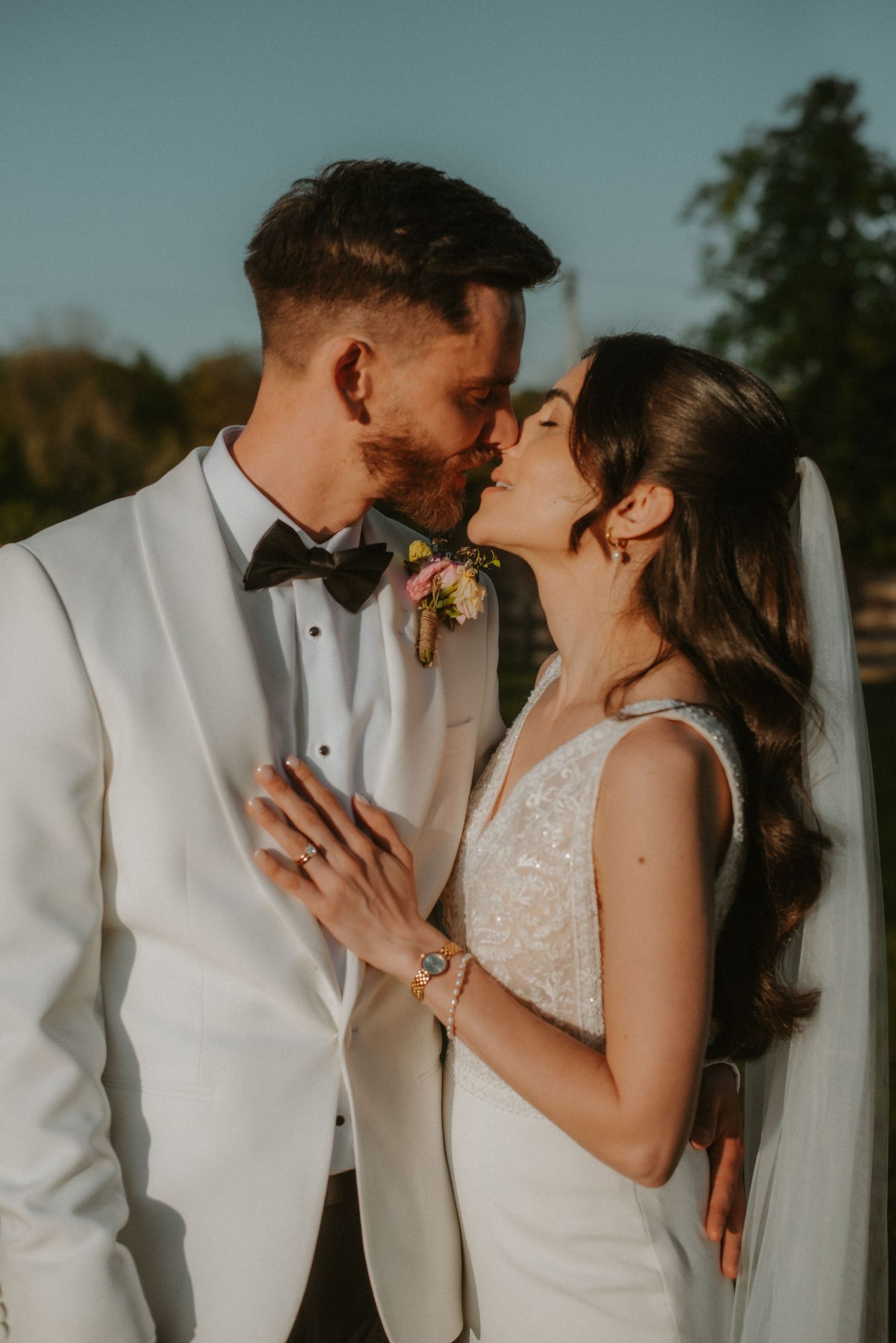 A bride and groom share a romantic kiss outdoors, with the groom in a white tuxedo and bride in a lace wedding gown.