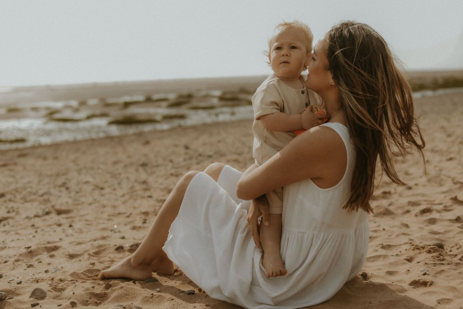 A mother in a white dress sits on a sandy beach holding her toddler, sharing a tender moment by the ocean.