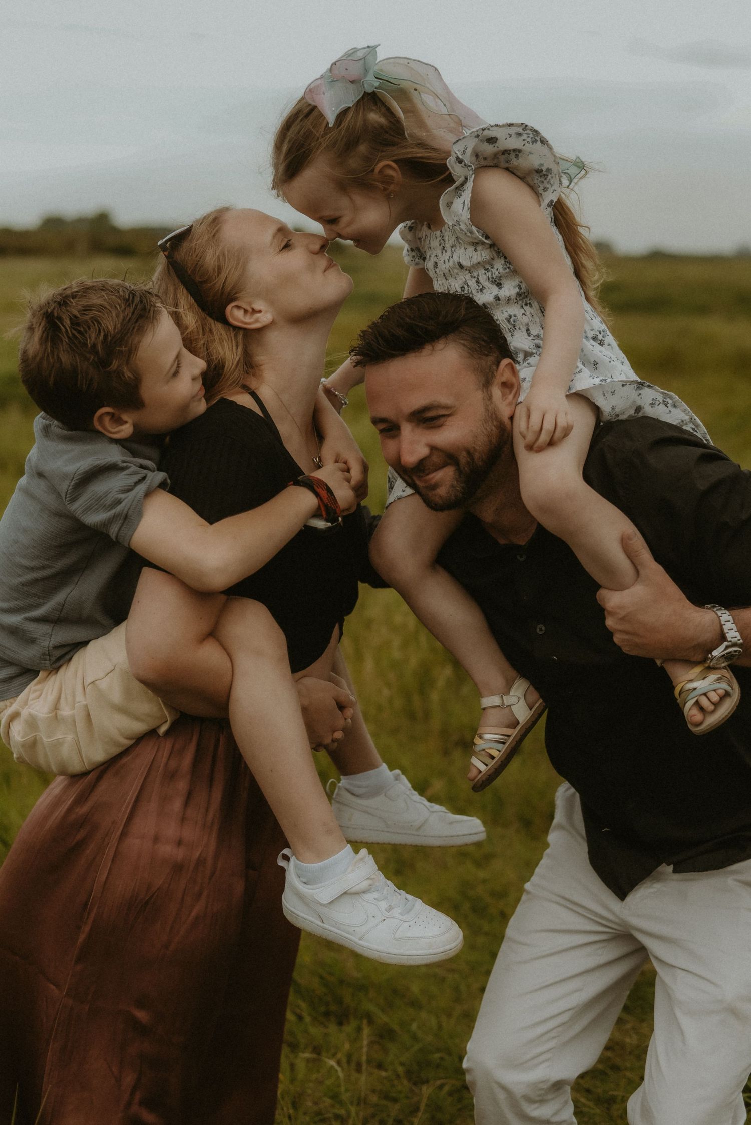 Happy family of four playing outdoors in a green field, parents lifting children while sharing a joyful kiss.
