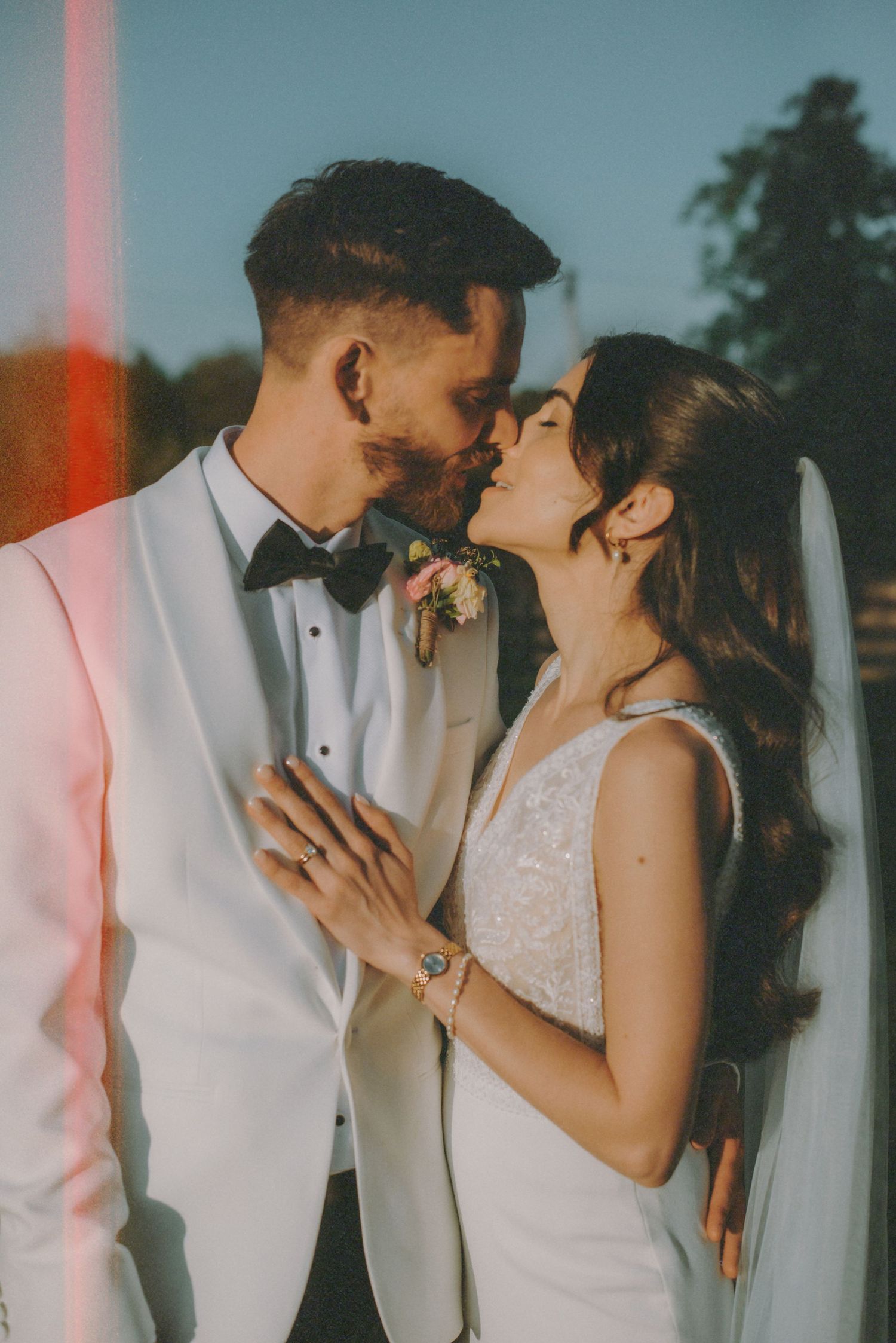 A bride and groom share a romantic kiss in matching white wedding attire outdoors on their wedding day.