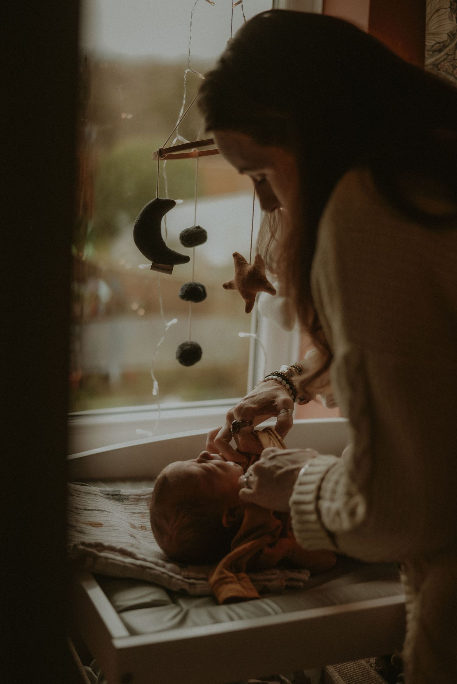 A mother tenderly interacts with her newborn baby by a window with a moon and star mobile hanging above.