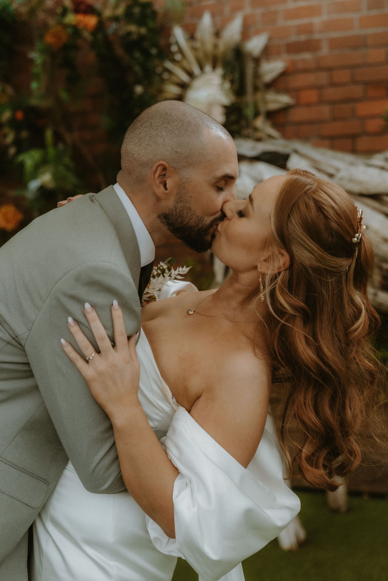 A bride and groom share a romantic kiss in wedding attire, with a brick wall and lush greenery backdrop.