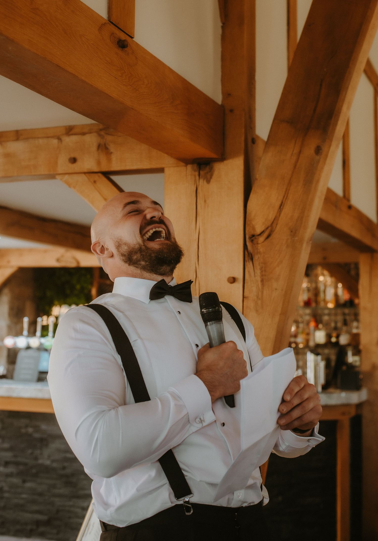 Bald groom laughing joyfully while giving a wedding speech, holding microphone and paper, wearing white shirt with suspenders and bow tie.