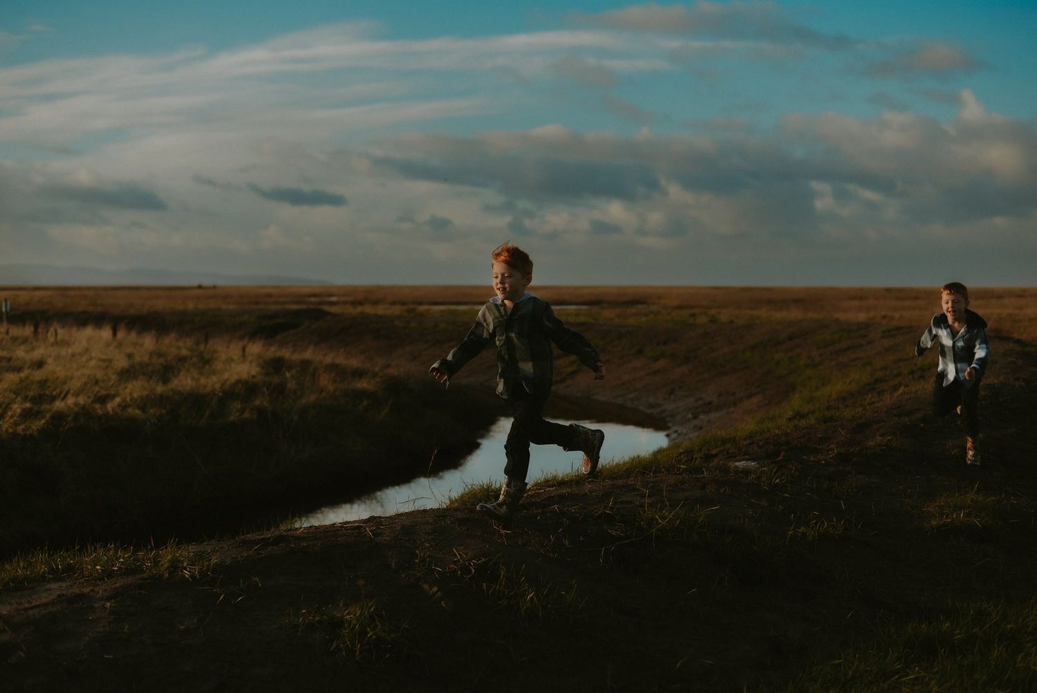 Child running across vast open field beside stream under dramatic cloudy sky at dusk.