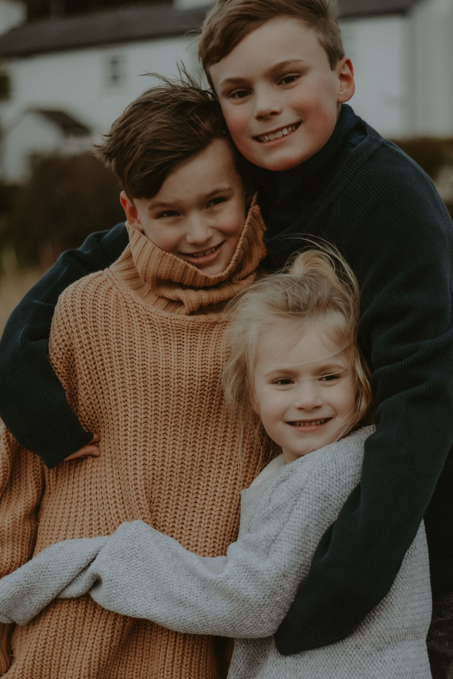 Three smiling siblings hugging closely outdoors wearing cozy knit sweaters in warm autumn light.