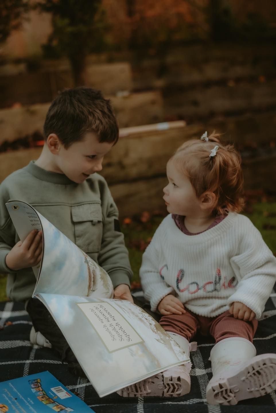 Two young siblings sit outdoors on a blanket, sharing a picture book together in autumn light.