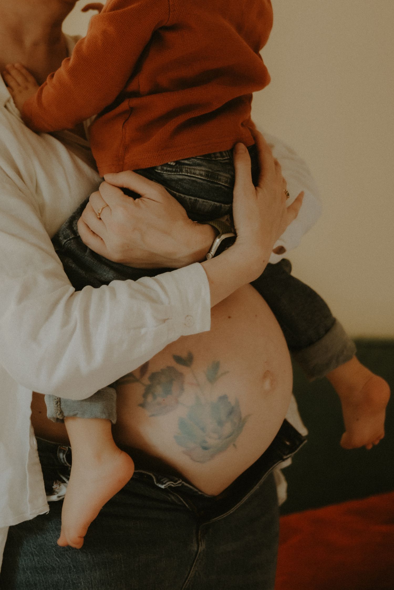 Pregnant woman in white shirt with floral tattoo on belly holding toddler in rust hoodie.