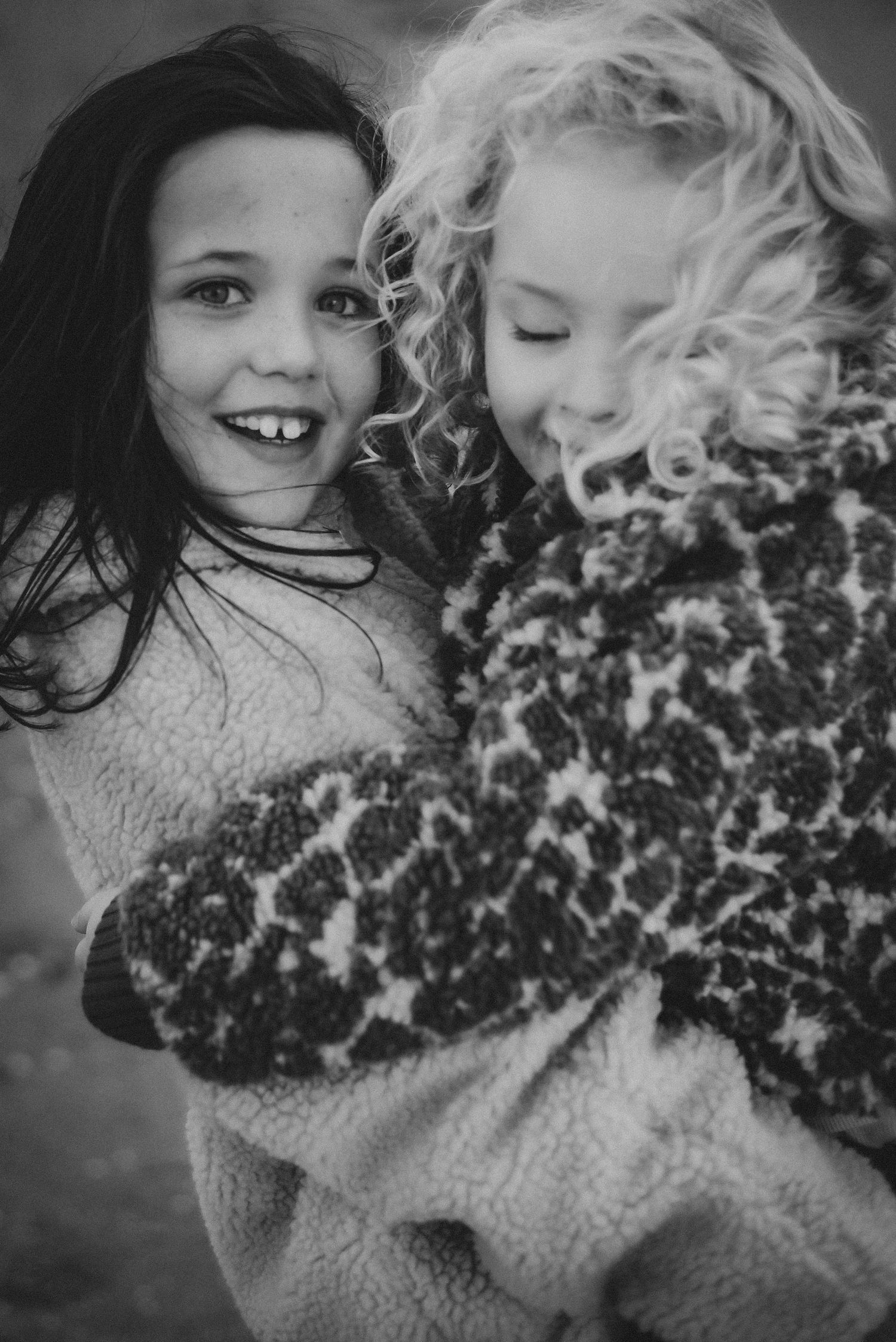 Black and white portrait of two young girls hugging and laughing together outdoors in cozy sweaters.