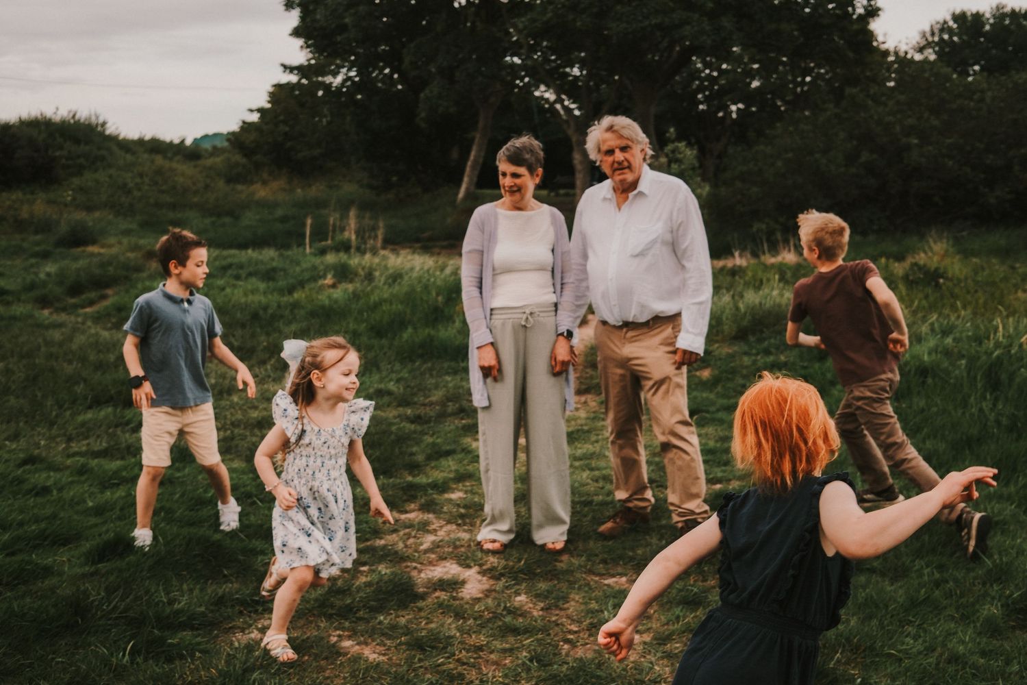 Family gathering outdoors on green grass, parents watching children play in a scenic countryside setting at golden hour.