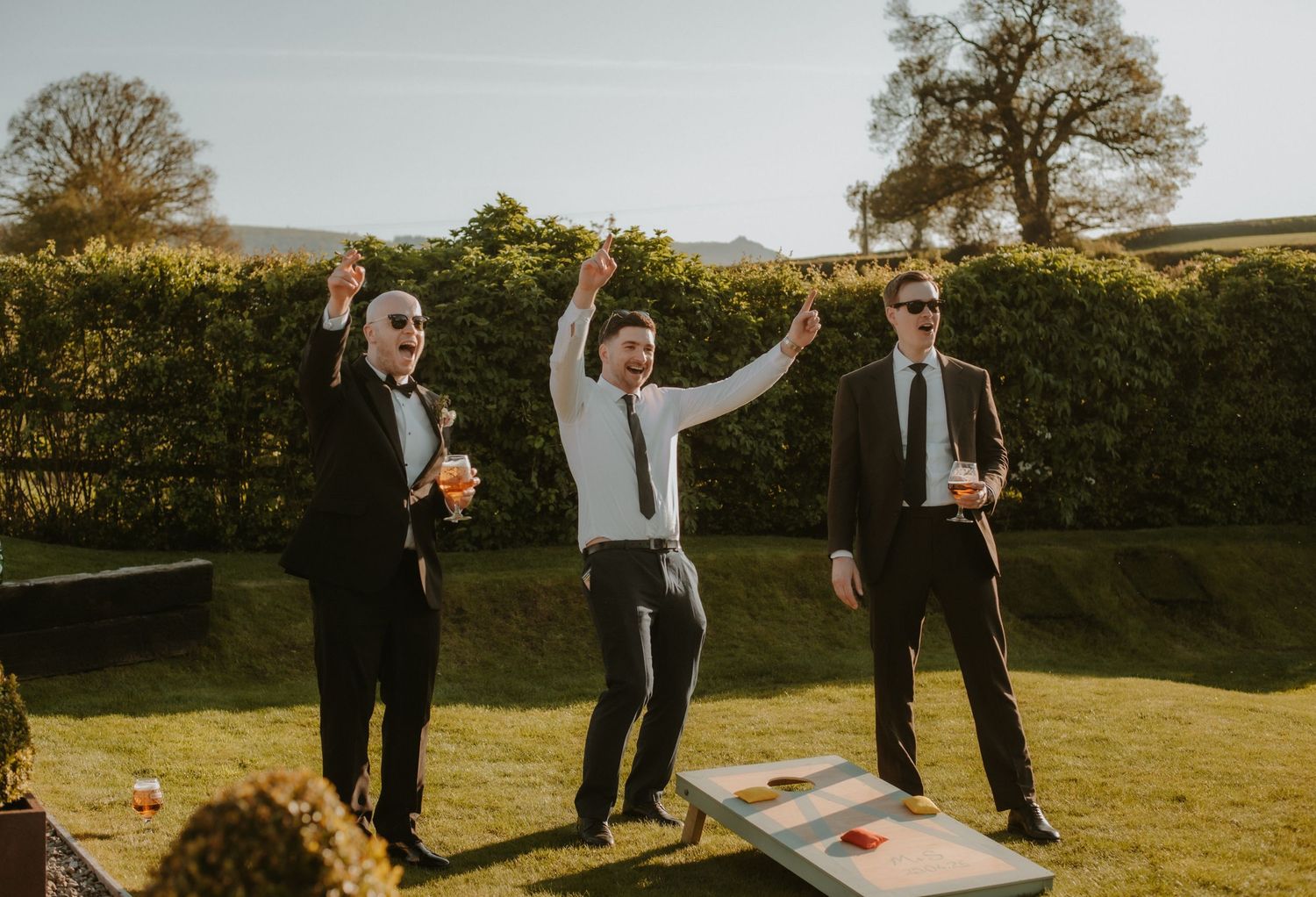 Three groomsmen celebrating outdoors playing cornhole lawn game at a sunny wedding reception.