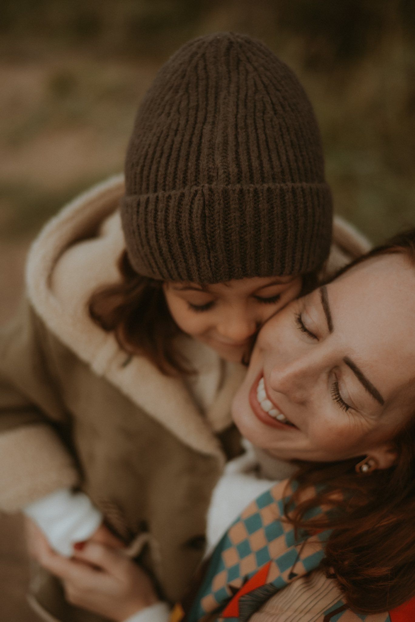 A smiling mother embraces her young child wearing a brown knit beanie outdoors in autumn.